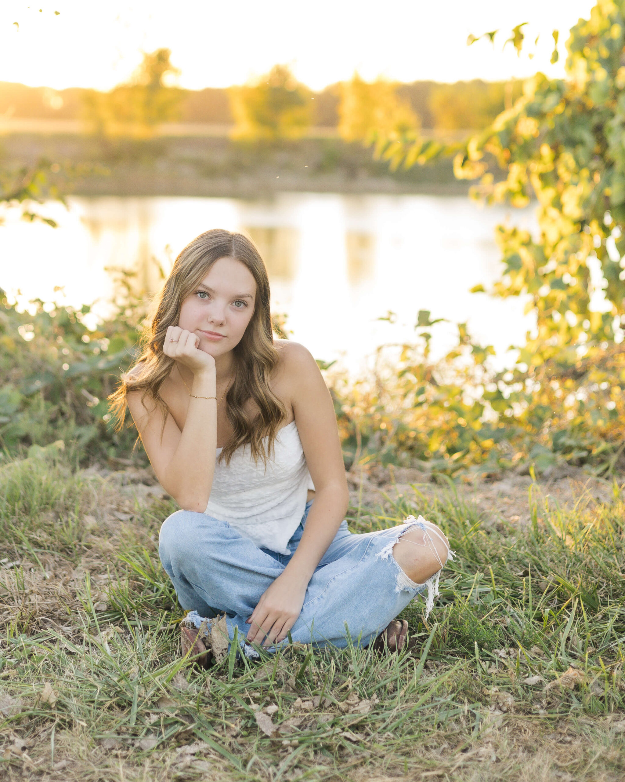 A high school senior sits cross legged by a lake at sunset in a white shirt leaning on her wrist after visiting tanning salons in Springfield, IL