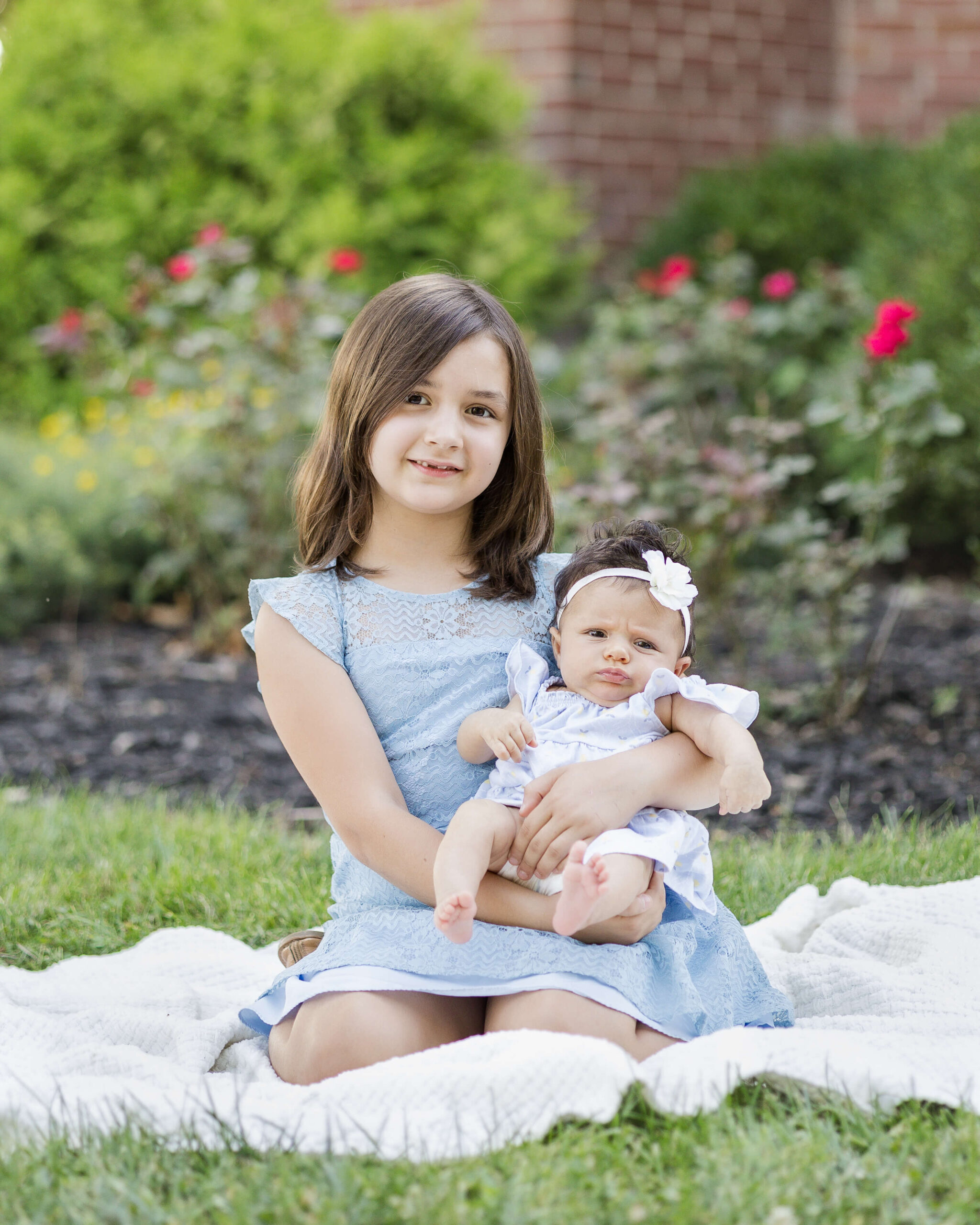 A toddler girl sits holding her baby sister in a garden on a picnic blanket after enjoying childcare in springfield, il