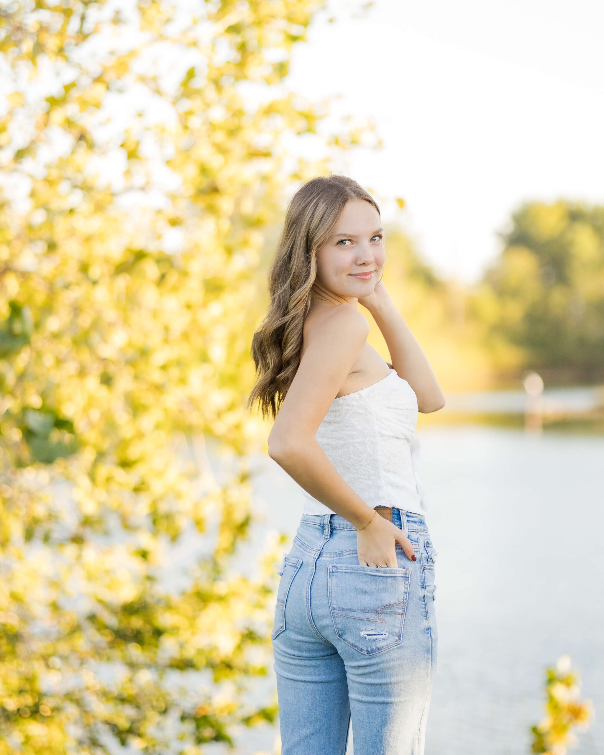 A teenager in a white shirt and jeans stands with a hand in her back pocket by a pond