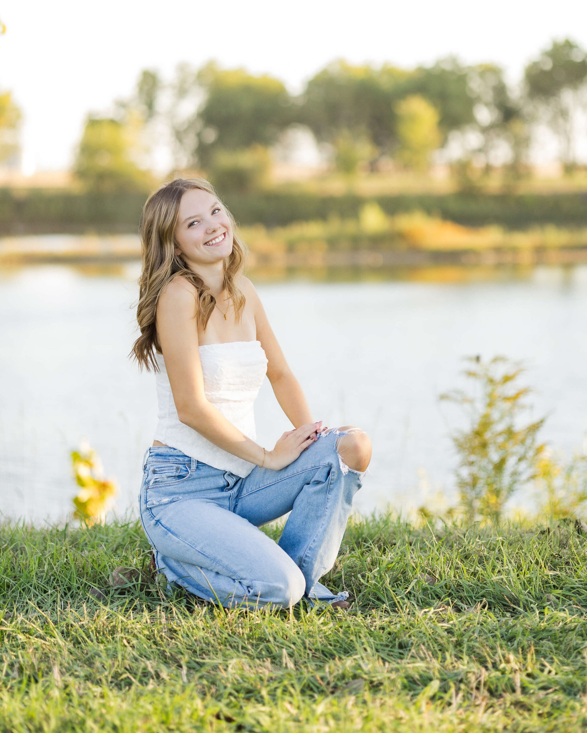 A high school senior kneels by a pond smiling in a white top and jeans after exploring graduation party venues in springfield il