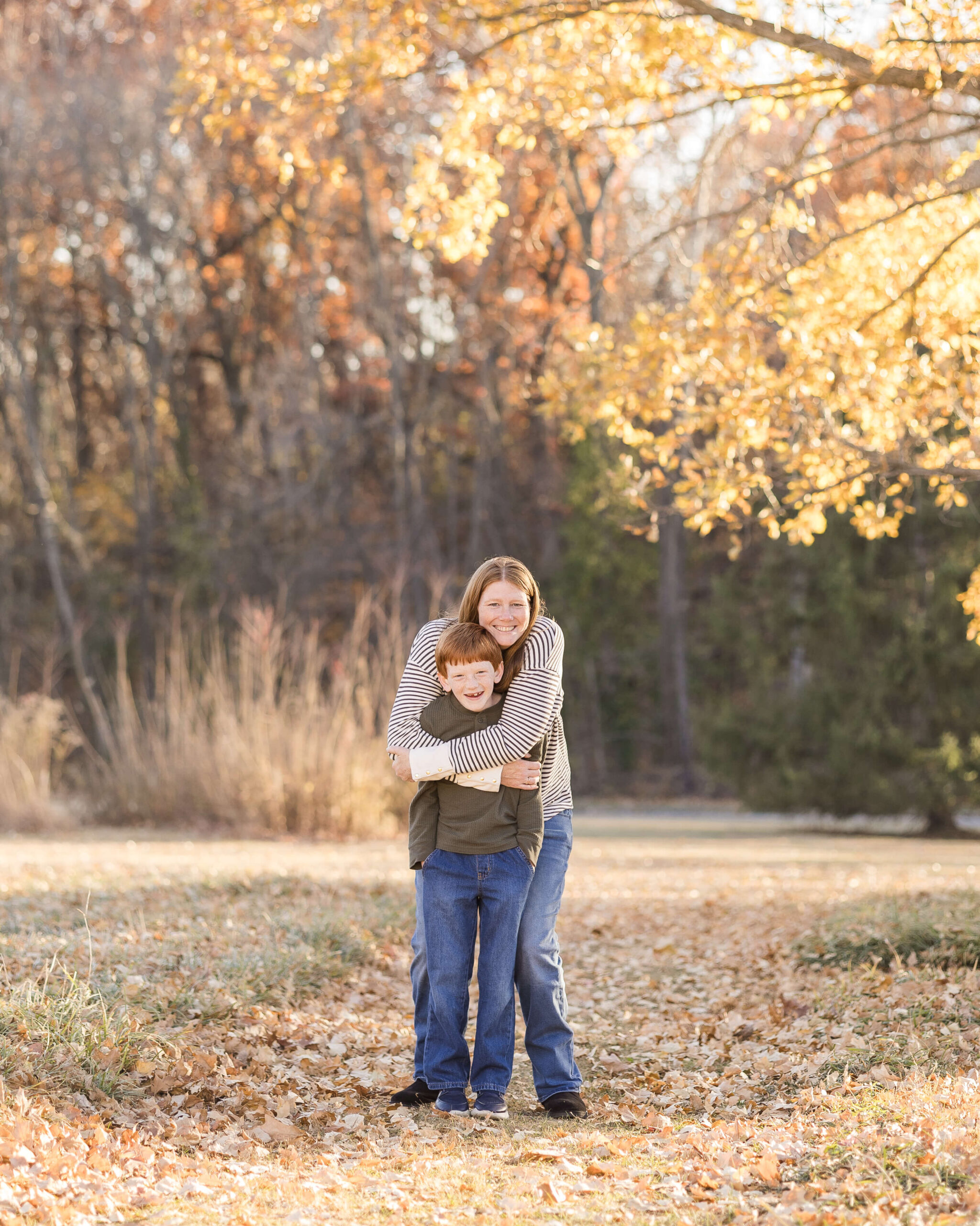 A happy mom in a stripe shirt hugs her young son in a park at sunset under a tree during fall