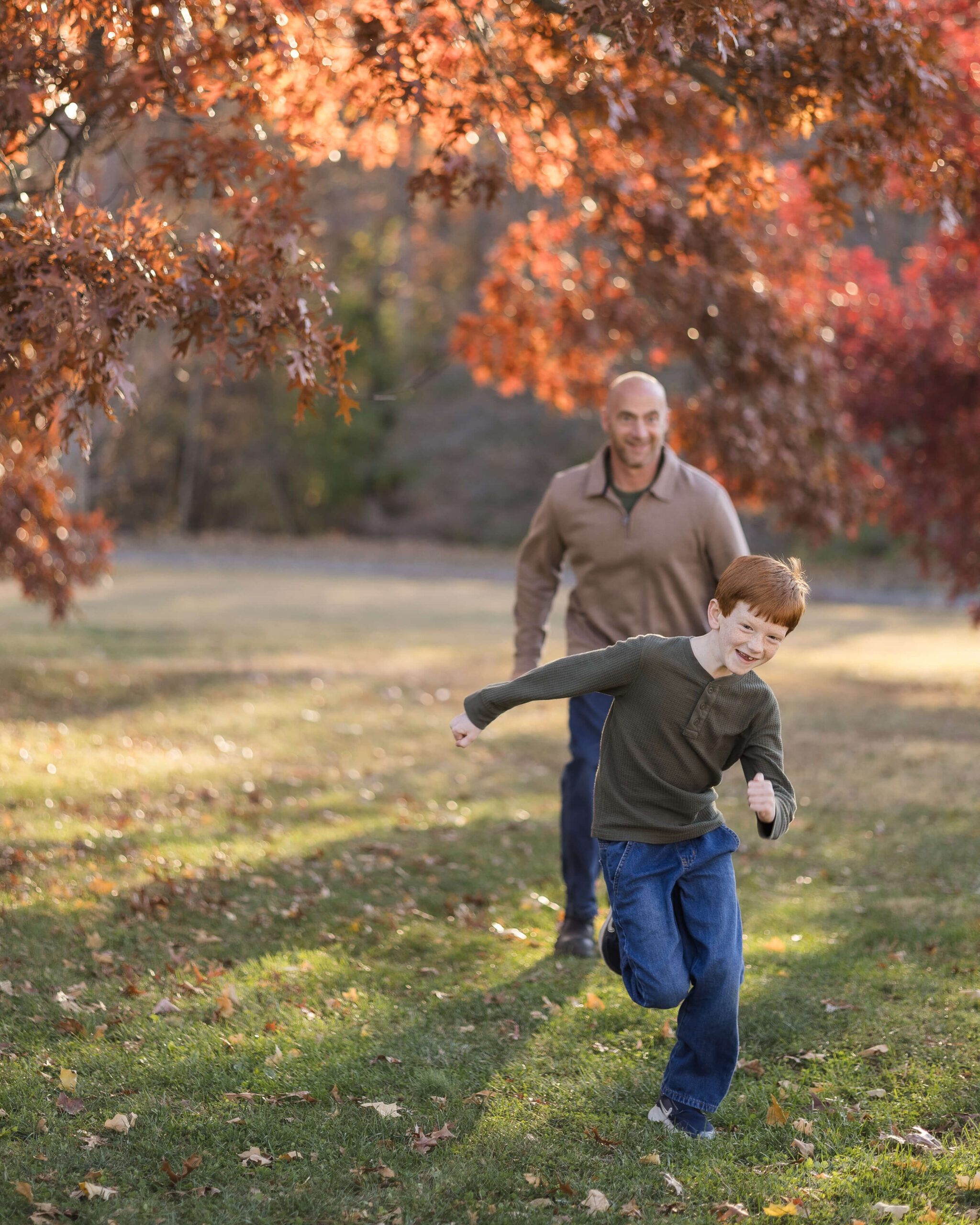 A young boy in a green shirt runs from dad in a park under trees in fall at sunset before heading to indoor playgrounds in springfield il