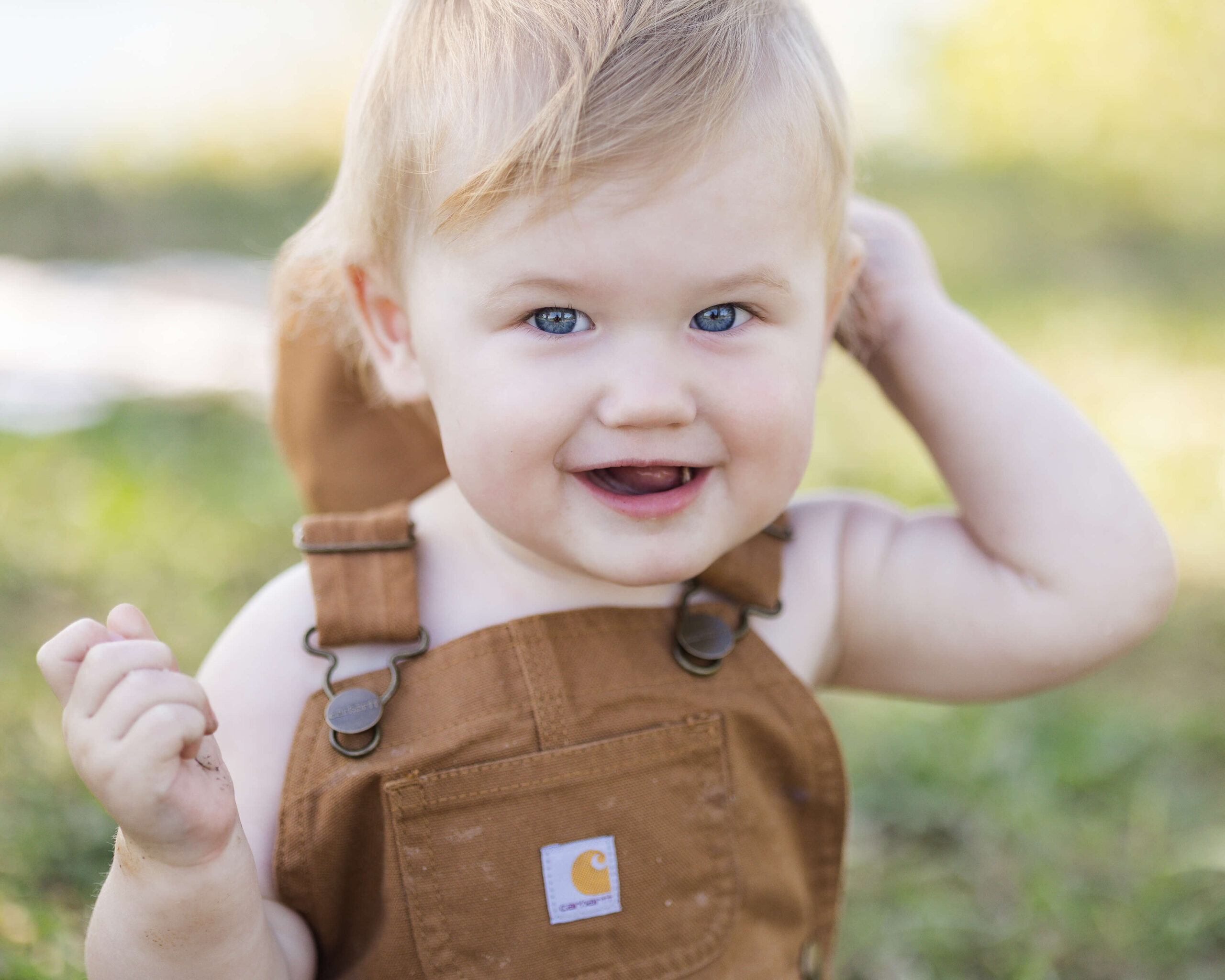 A closeup of a smiling toddler boy in brown overalls in a park lawn