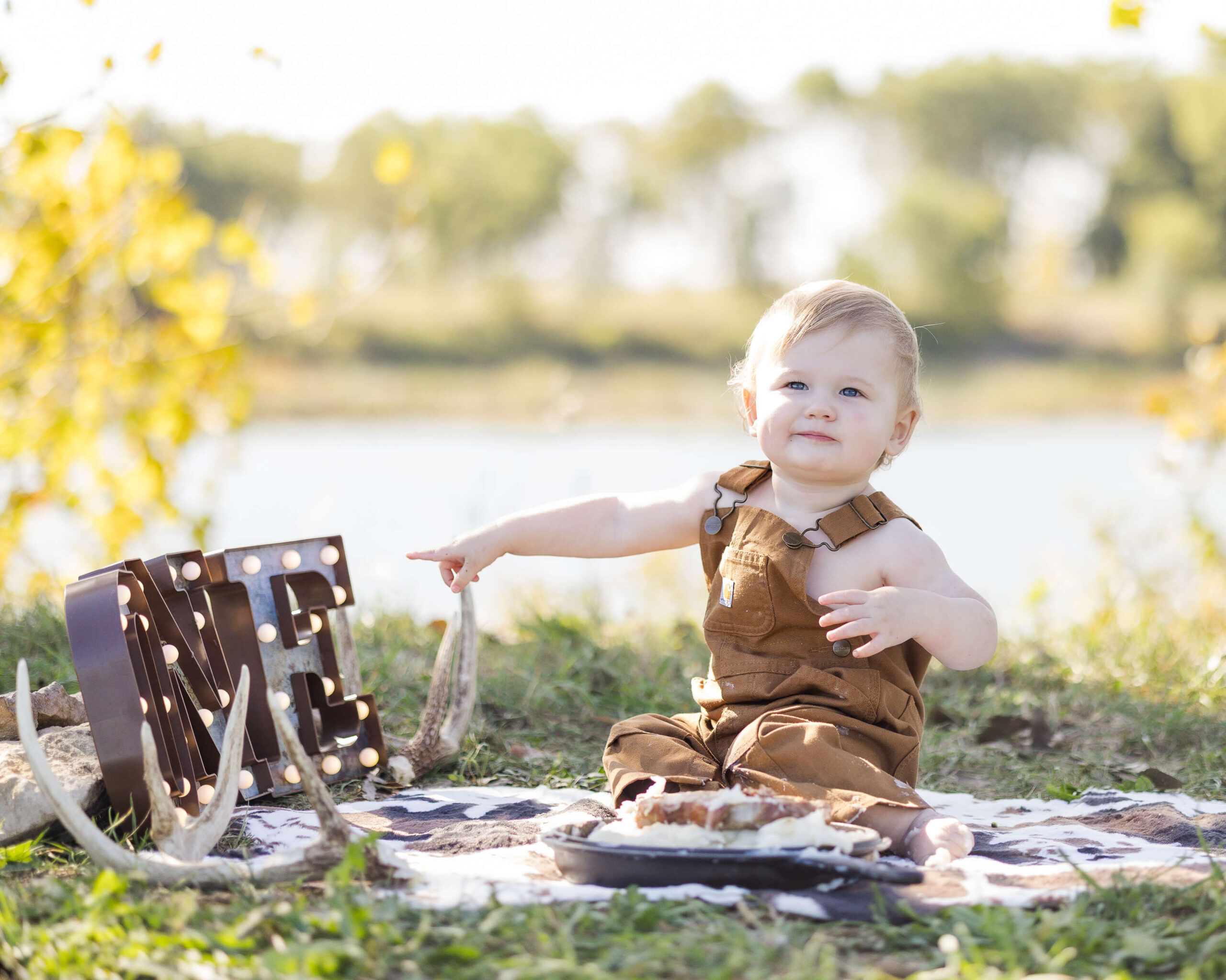 A toddler boy in brown overalls points to ONE in marquee letters in a park lawn by a pond after enjoying kid friendly hotels springfield il