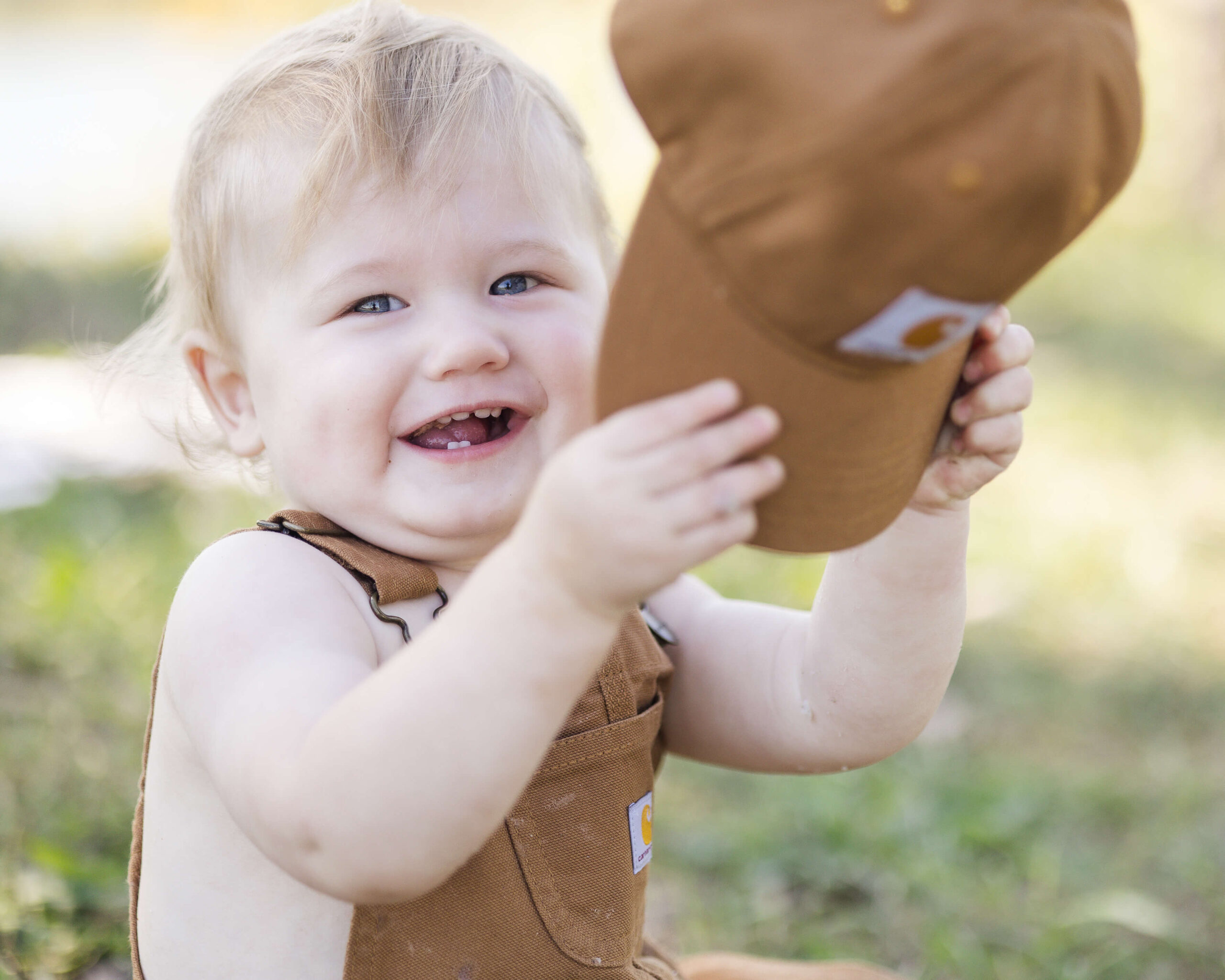 A young toddler in brown overalls plays with a matching hat in a park lawn with a big smile after visiting kid friendly hotels springfield il