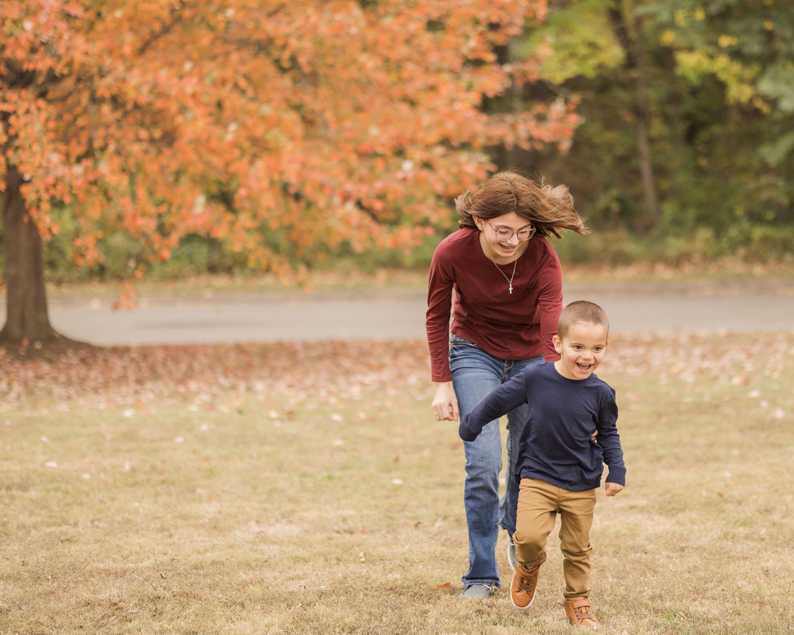 A teen girl chases her toddler brother in a park lawn in fall