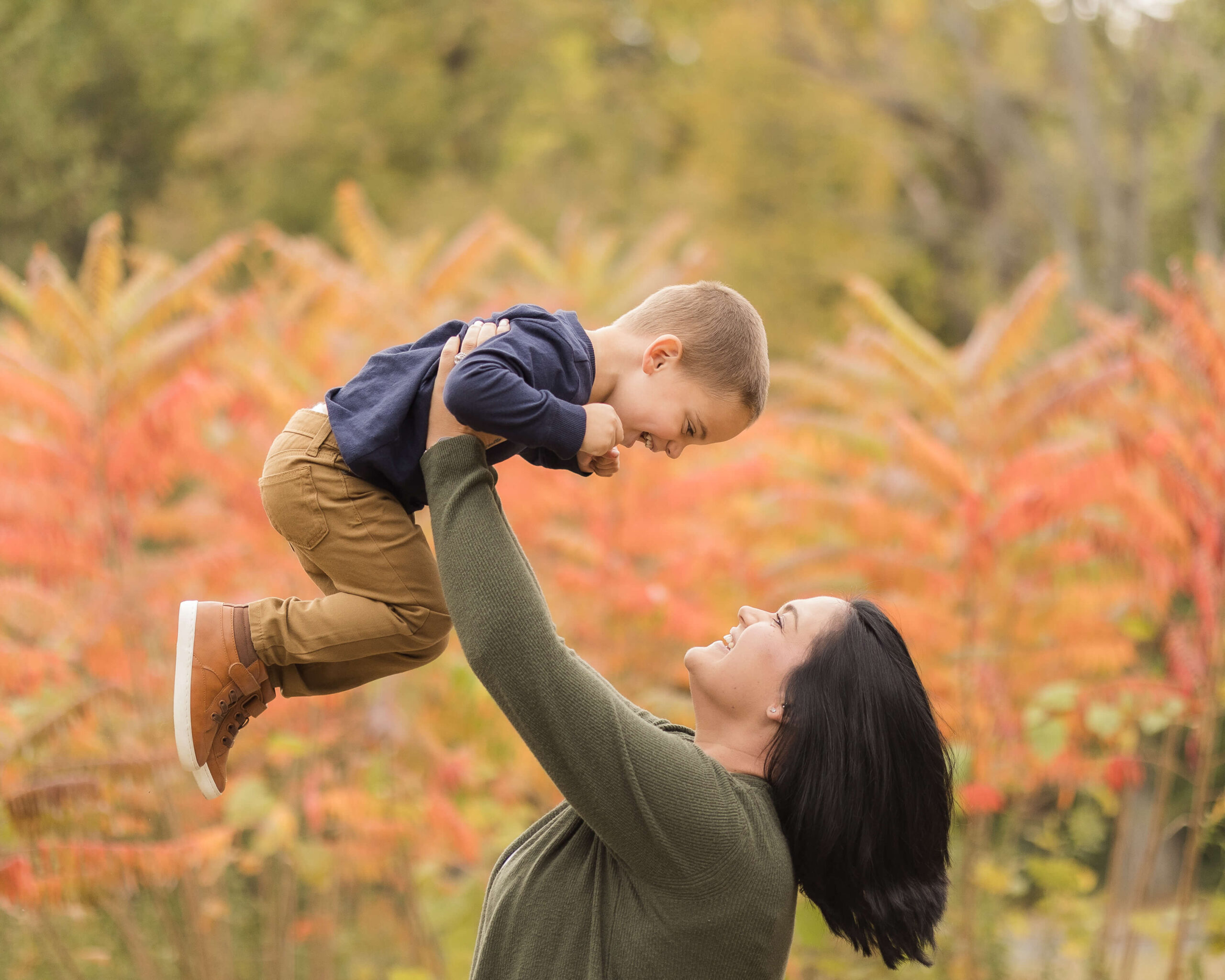 A happy mom lifts her giggling toddler son over her head in a park in fall after visiting kid friendly restaurants in springfield il