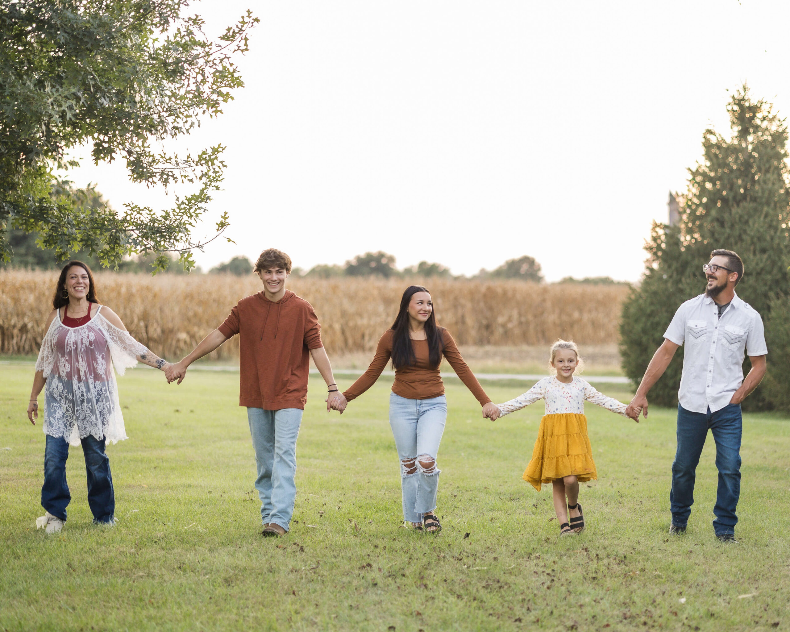 A mom and dad smile big while walking holding hands with their three kids in a park lawn at sunset