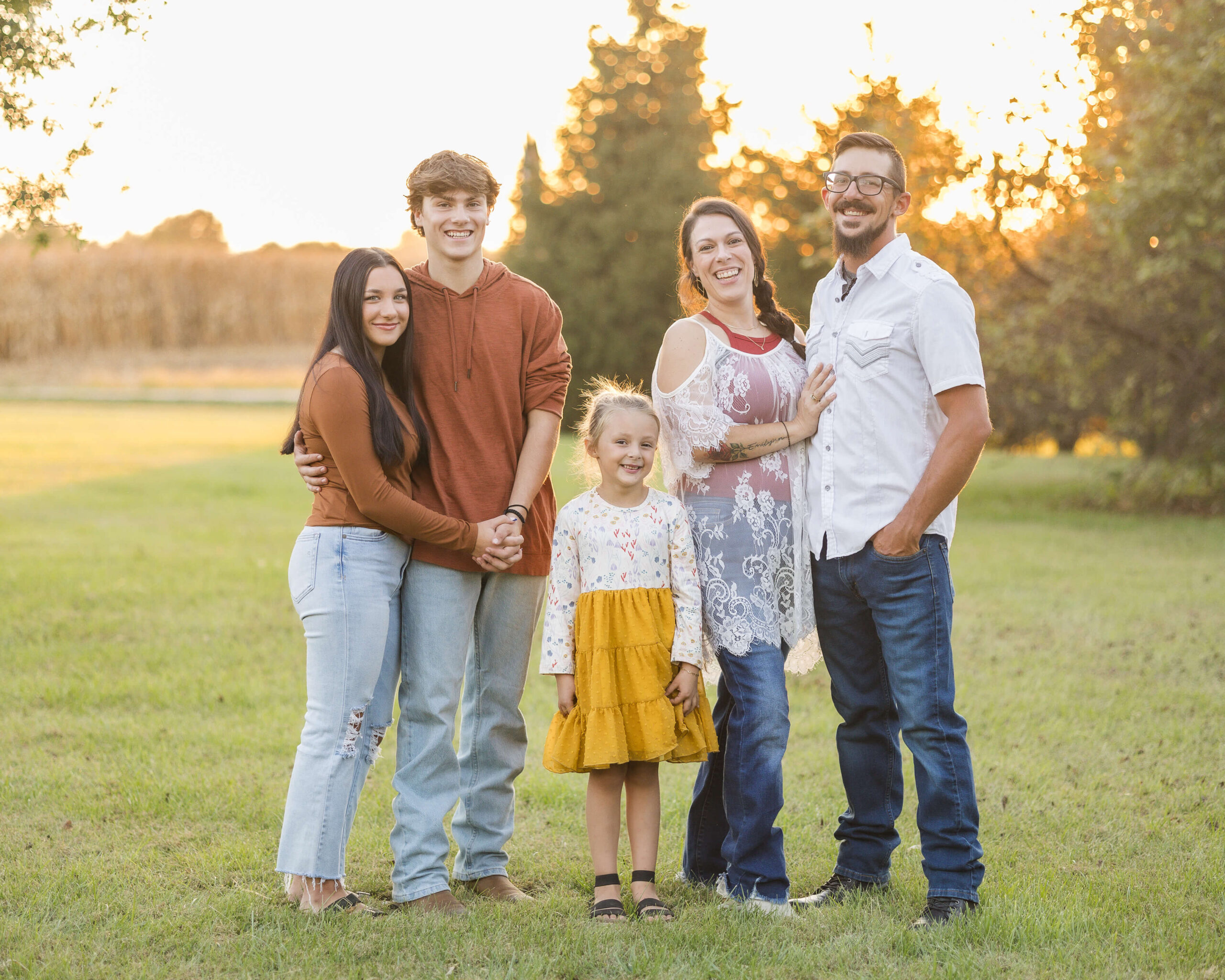 A happy family of 5 stand smiling in a park lawn at sunset in jeans after some parenting classes in Springfield, IL