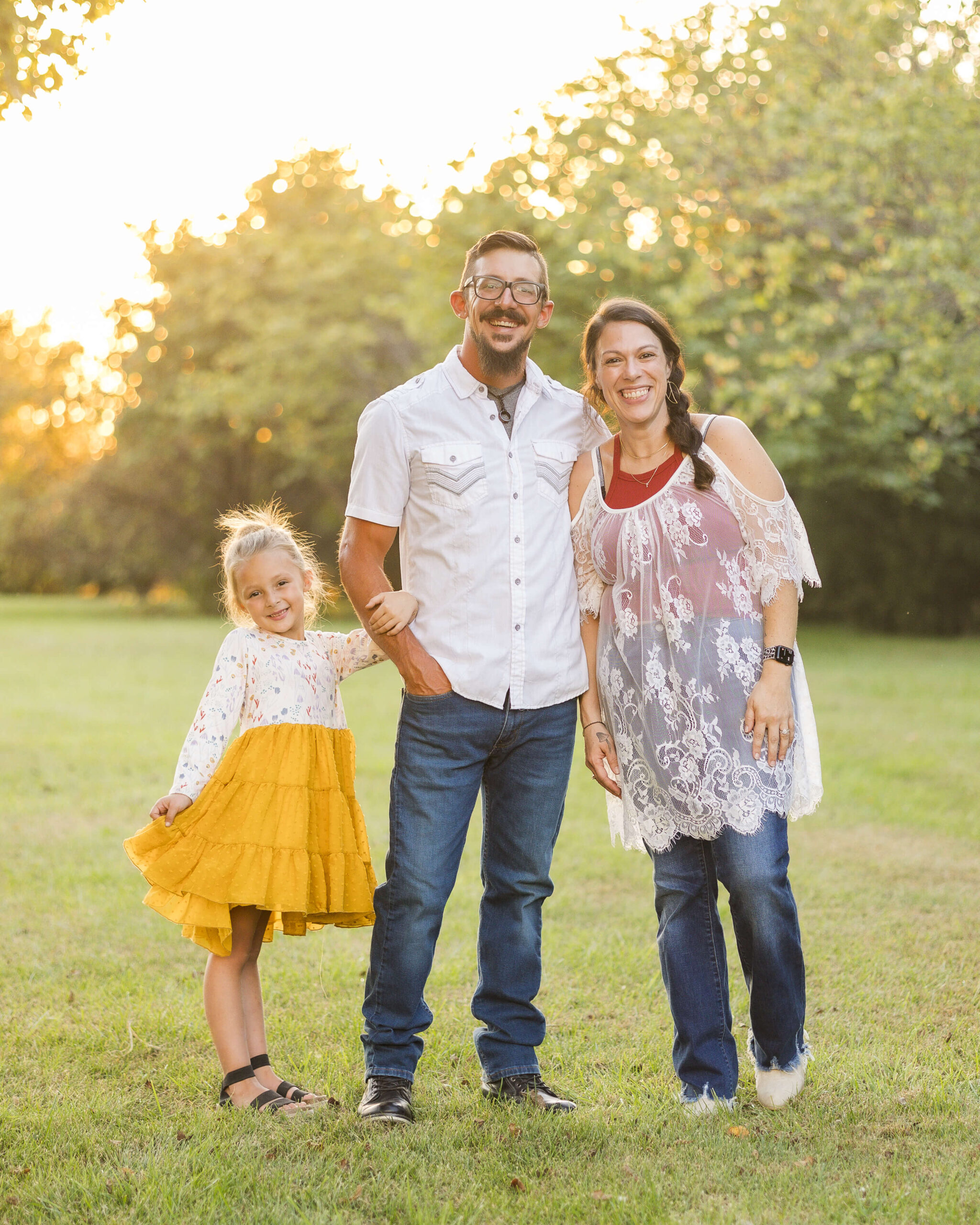 Happy mom and dad in jeans smile in a park lawn with their toddler daughter hanging on dad's arm in a yellow dress thanks to parenting classes in Springfield, IL