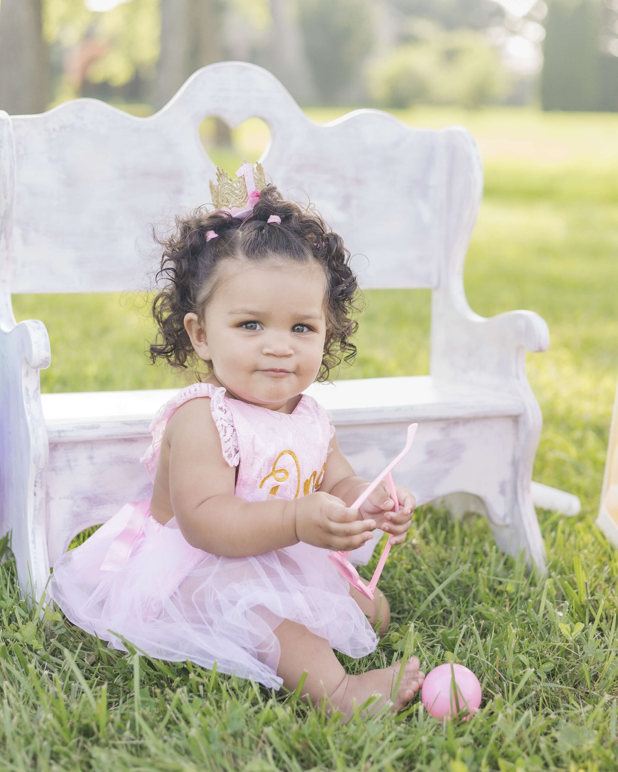 A toddler girl in a pink dress plays with matching pink toys in a lawn in front of a wooden bench at sunset