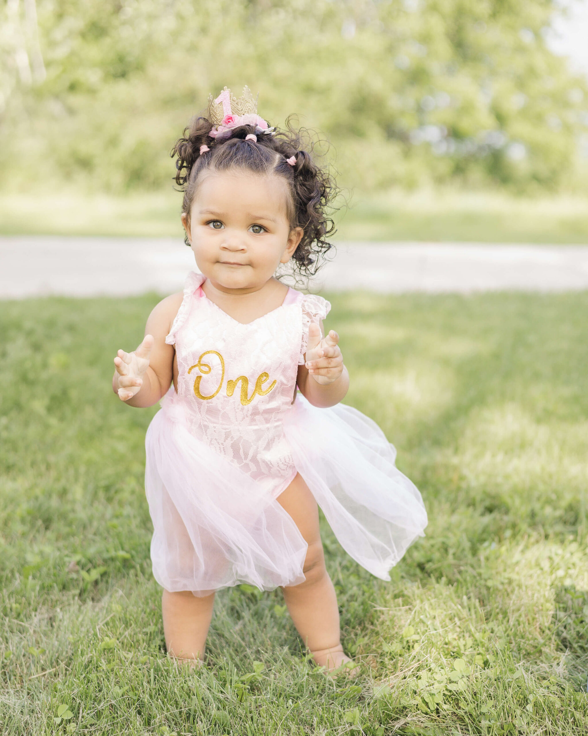 A happy toddler girl walks in some grass in a first birthday pink dress before visiting splash pads in springfield, IL