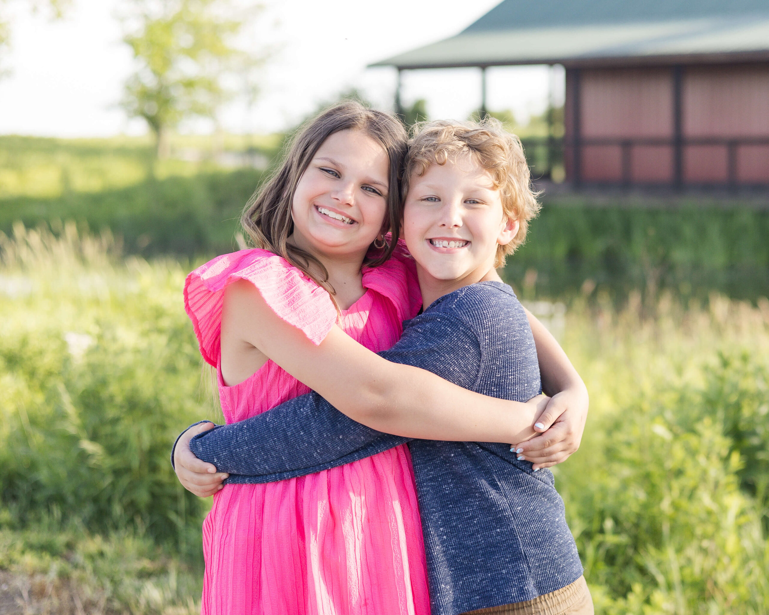 Happy toddler brother and sister hug while standing in pink and blue in a meadow at sunset