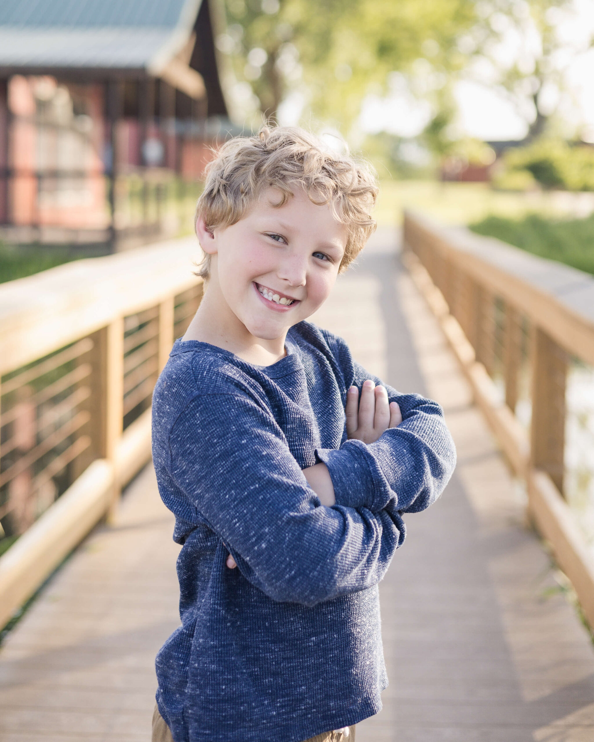 A happy toddler in a blue shirt crosses his arms and smiles while walking on a wooden bridge before heading to spring break camps springfield, IL