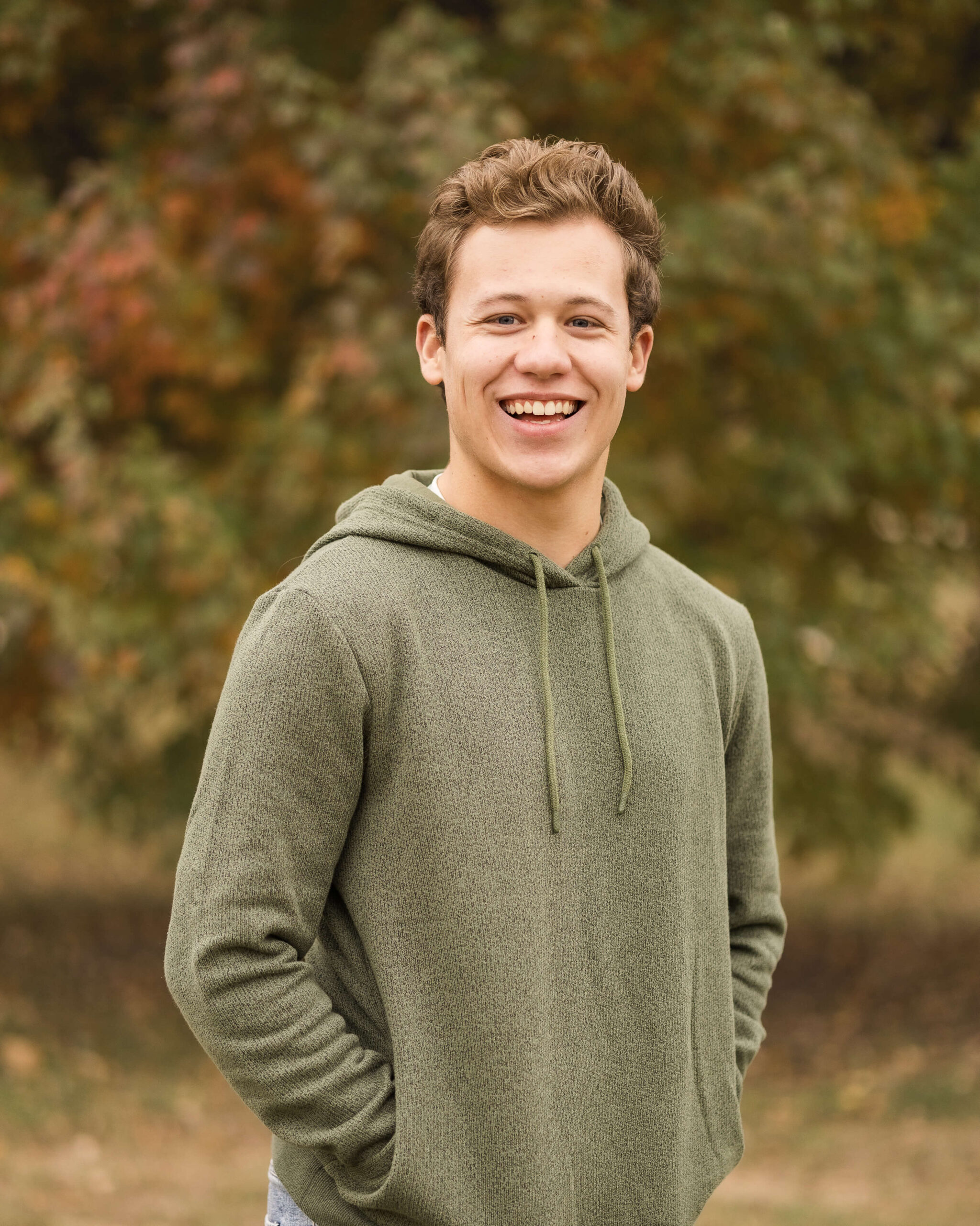 A happy high school senior stands smiling in a green hoodie with hands in pockets in a park