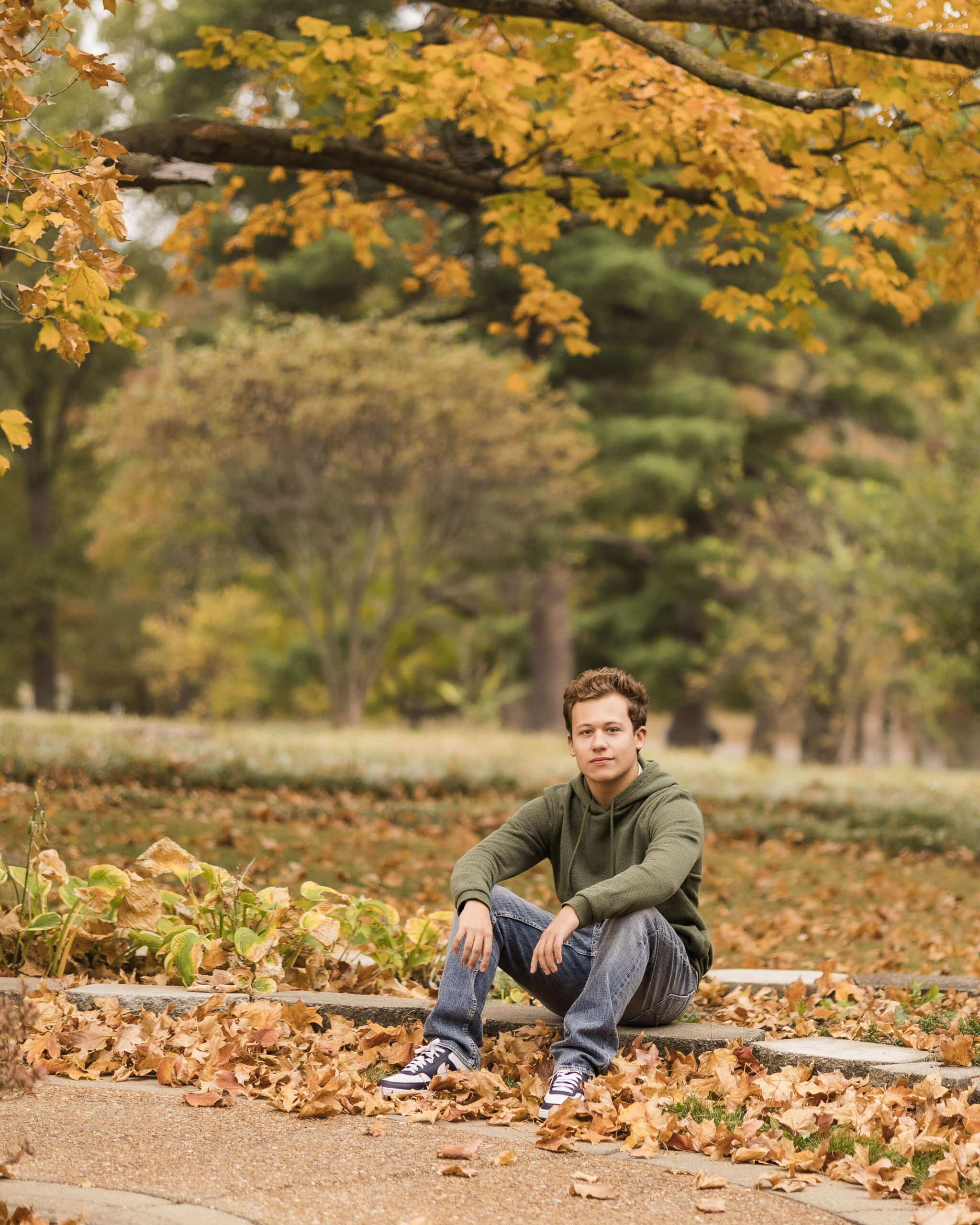 A teenage boy in a green hoodie sits on a small stone wwall in a park in fall after enjoying test prep in springfield il