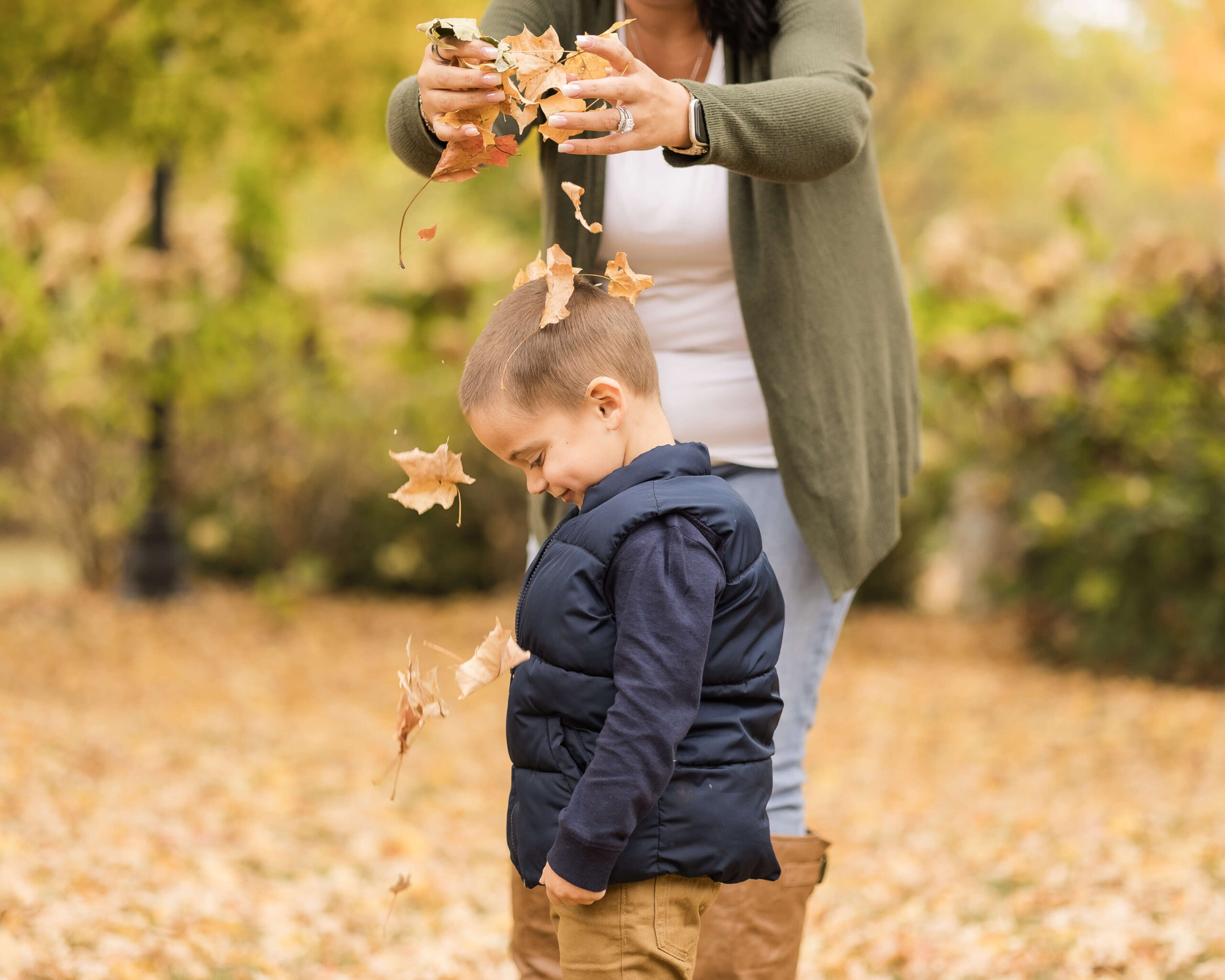A toddler boy stands in a blue sweater in a park as mom drops leaves over his head before visiting a trampoline park in springfield il