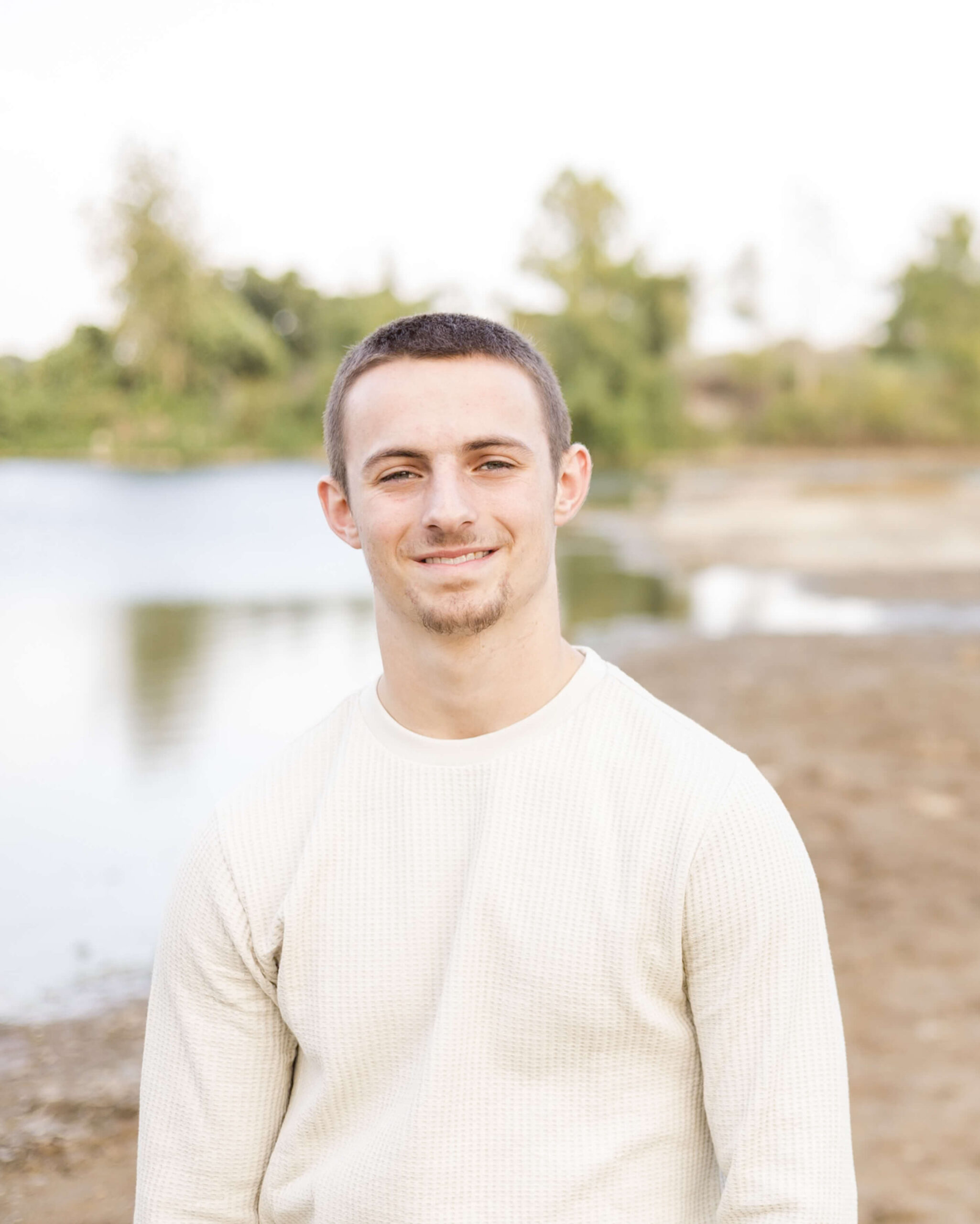 A smiling high school senior in a cream shirt next to a pond