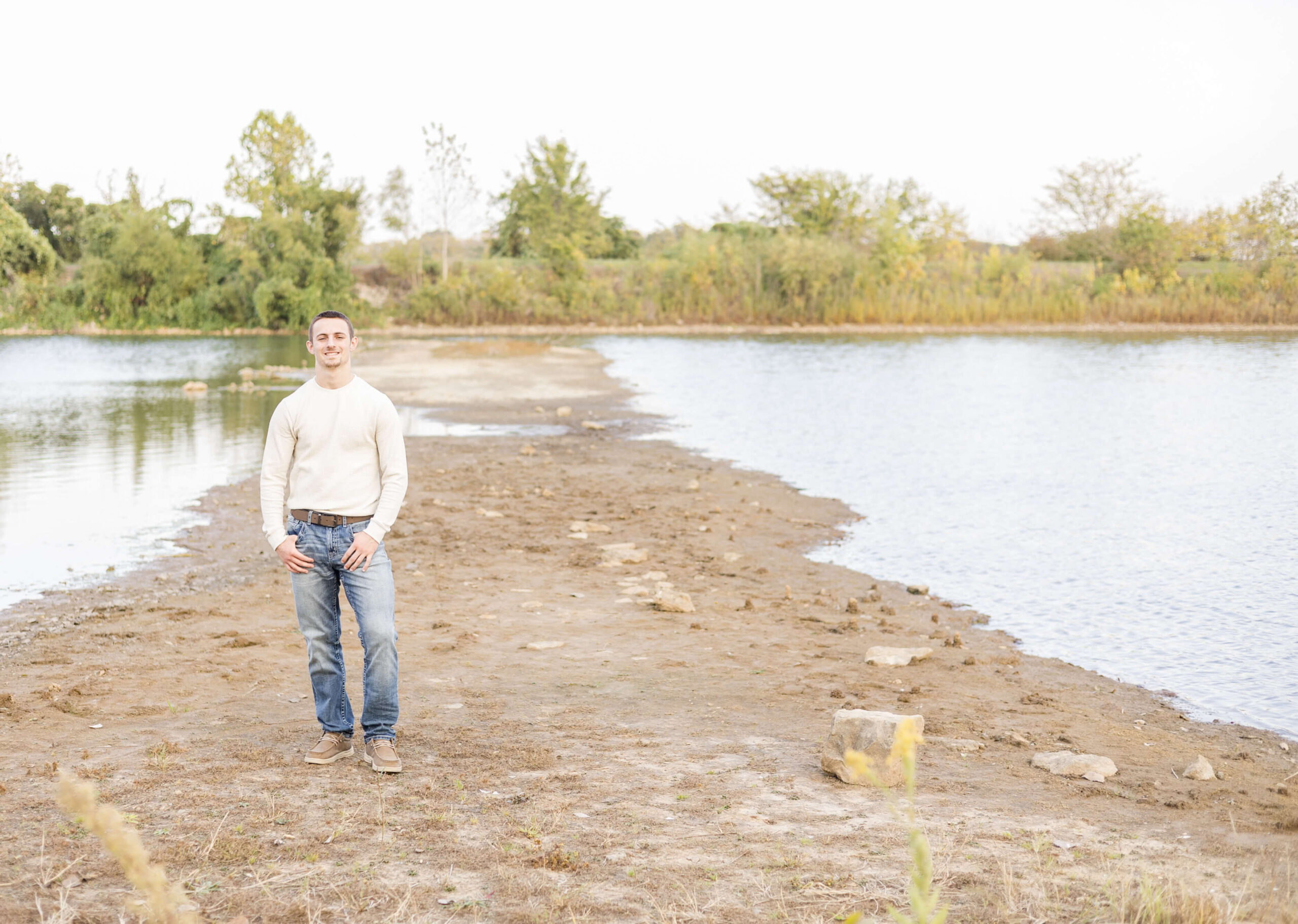 A high school senior in a white shirt and jeans stands on a sandbar in a pond at sunset smiling after some tutoring in springfield il