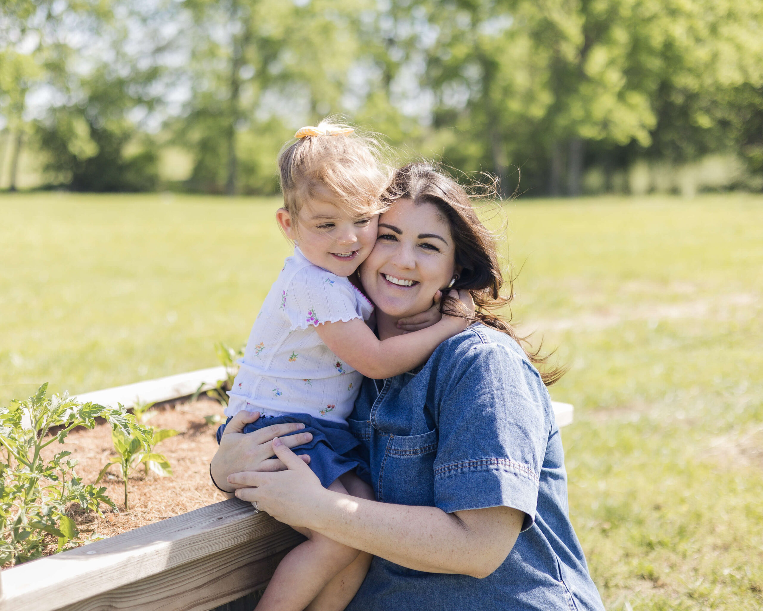 A toddler girl hugs her mom in a denim dress while sitting on a raised garden bed in a field