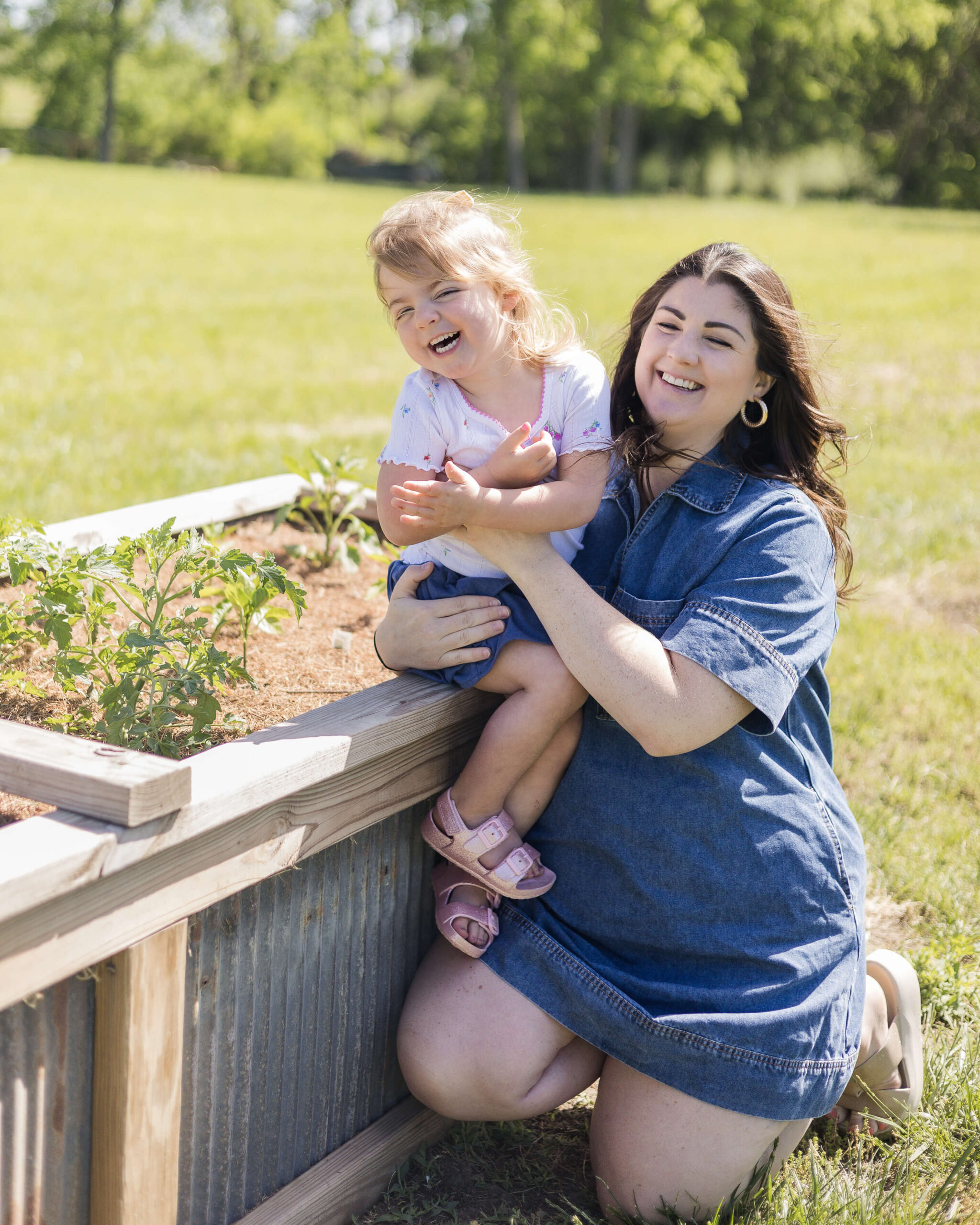 A mom and her toddler daughter giggle while kneeling and sitting with a raised garden in a field after finding birthday party places in Springfield, IL