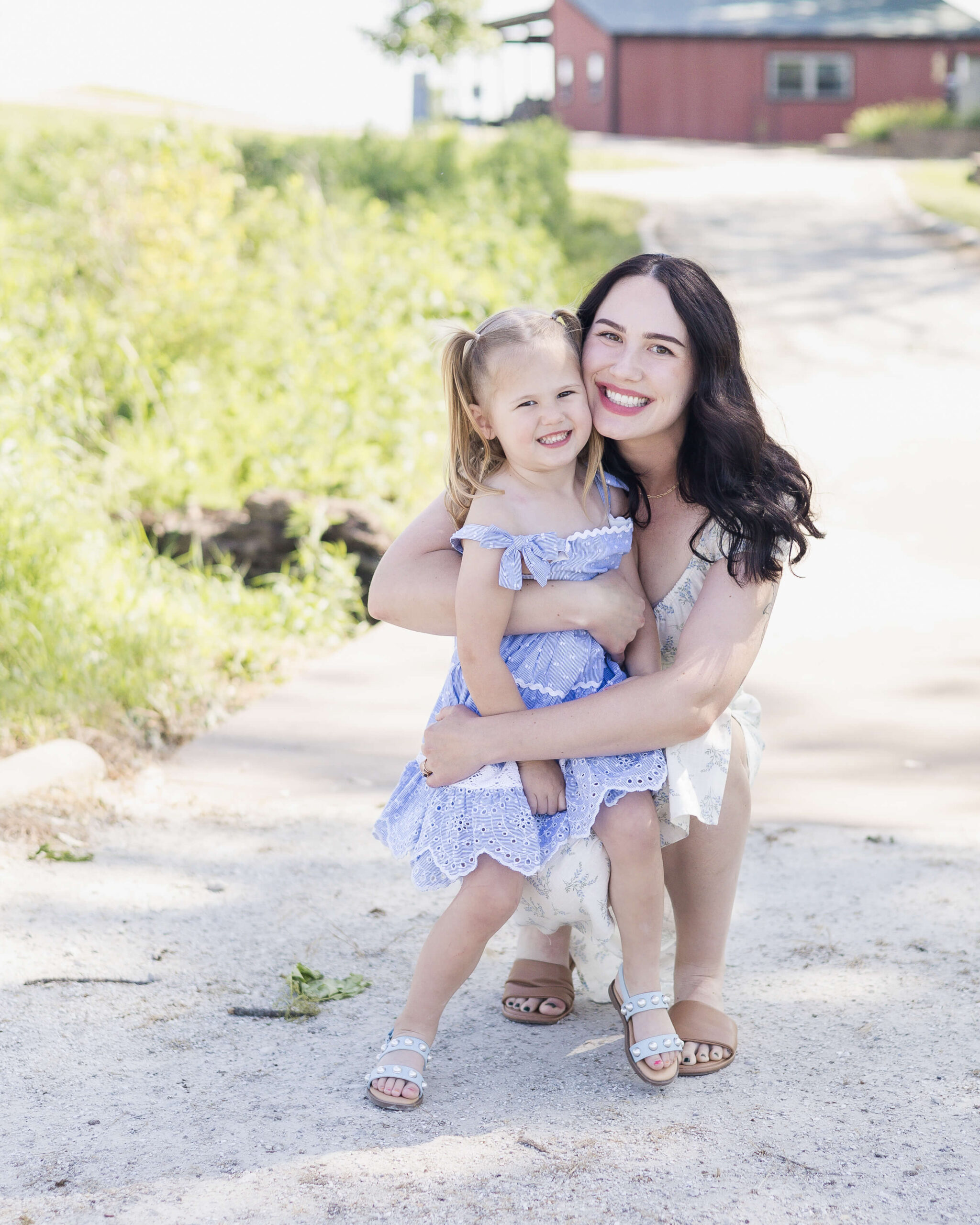 A happy mother in a floral print dress hugs her happy toddler daugher in blue in a farm trail