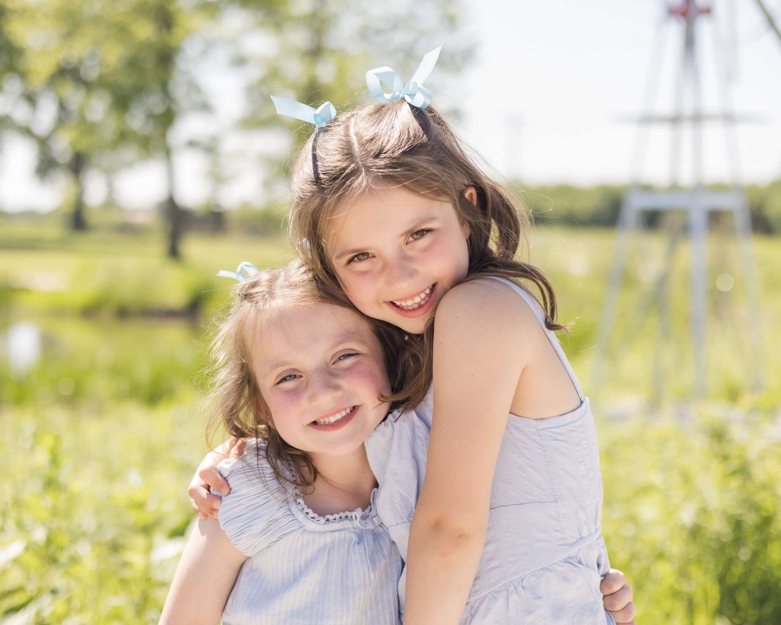 A toddler girl in a blue dress hugs her giggling little sister in matching dress in a park