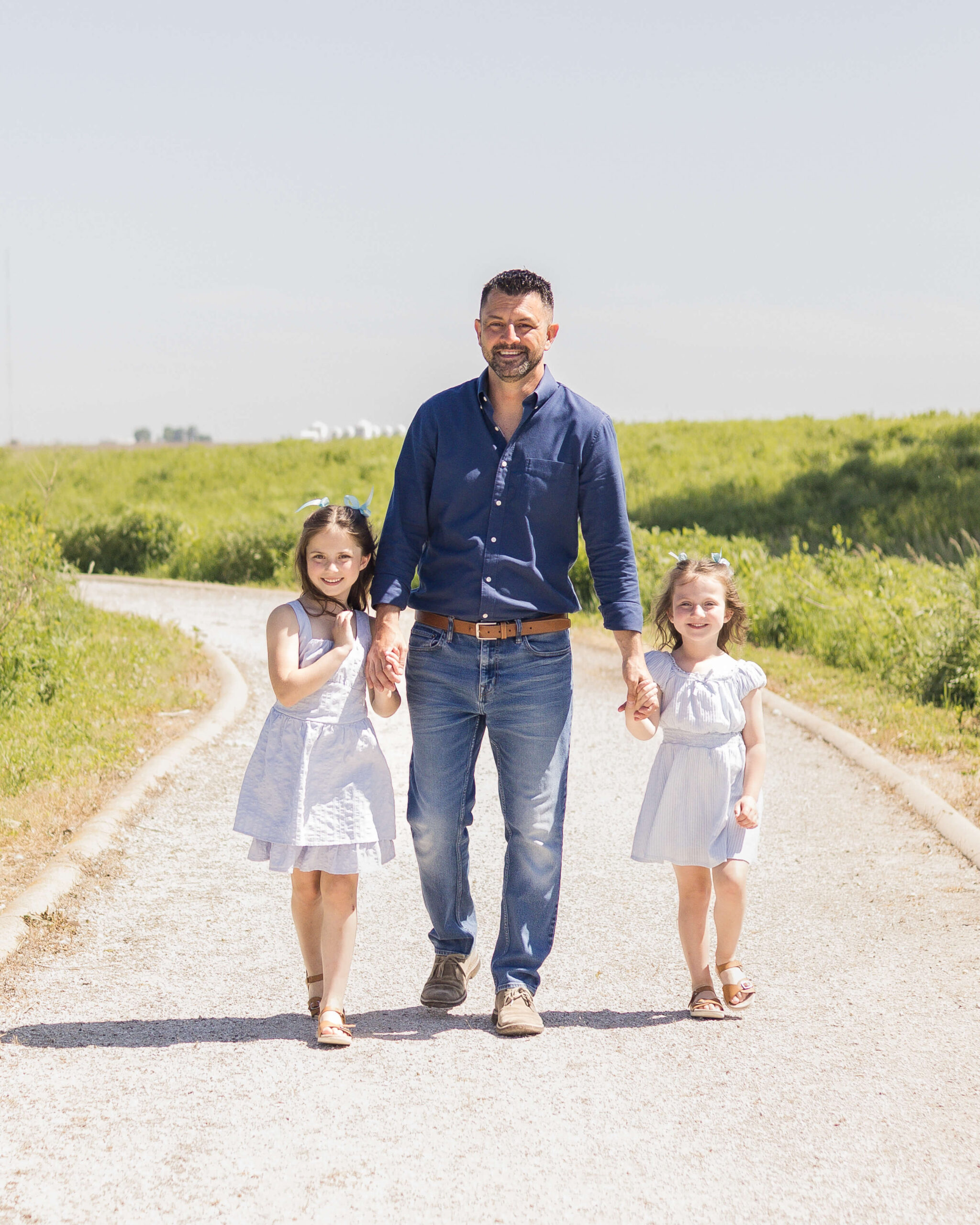 A dad in blue walks in a park path holding hands with his two toddler daughters in matching blue dresses after enjoying easter egg hunts in springfield il