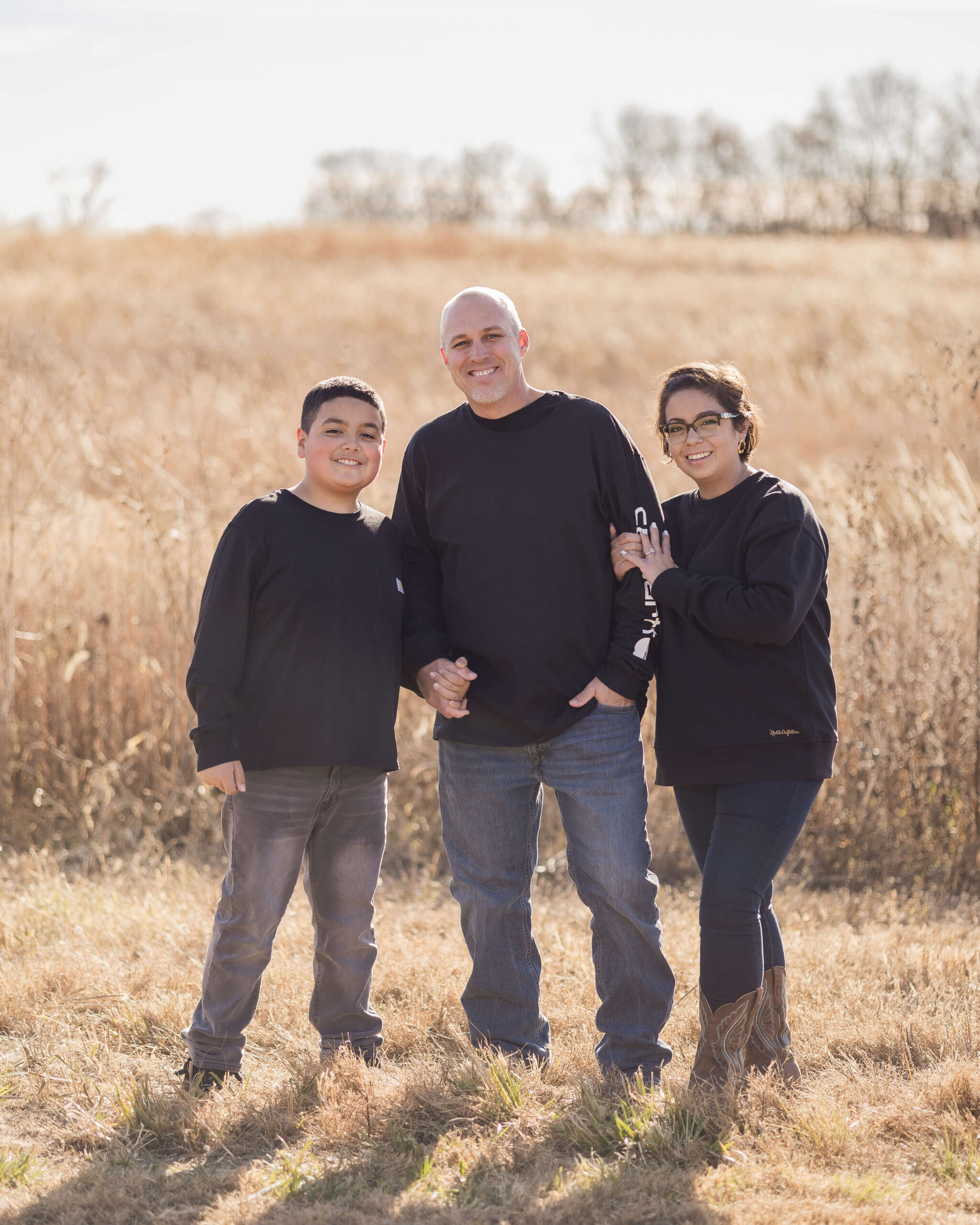 A happy teen boy wearing matching black long sleeve shirts stands with his mom and dad holding hands by a field of tall golden grass at sunset thanks to family counseling in springfield il