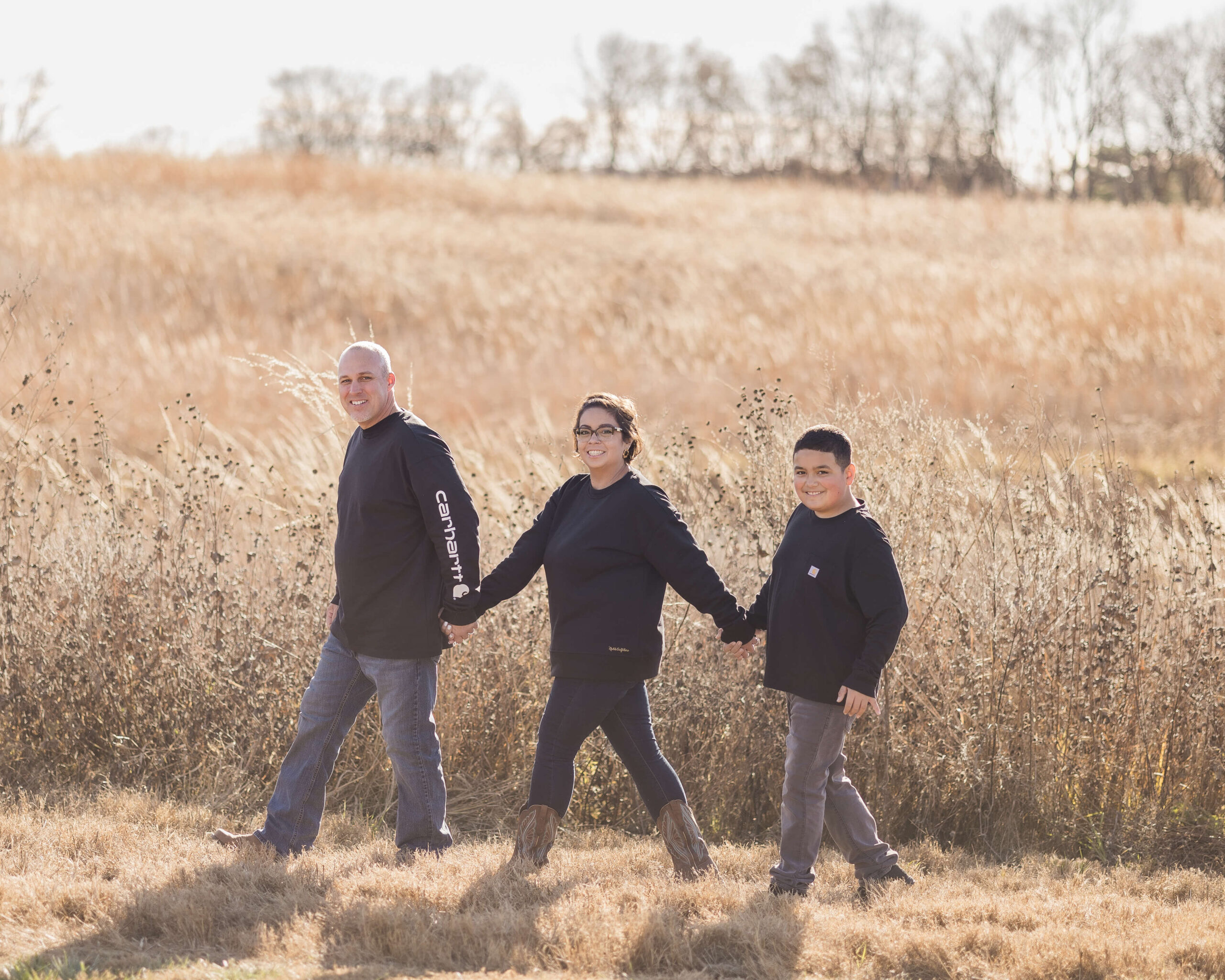 A mom and dad in black walk along a field of tall golden grains at sunset holding hands with their young son after some family counseling in springfield il