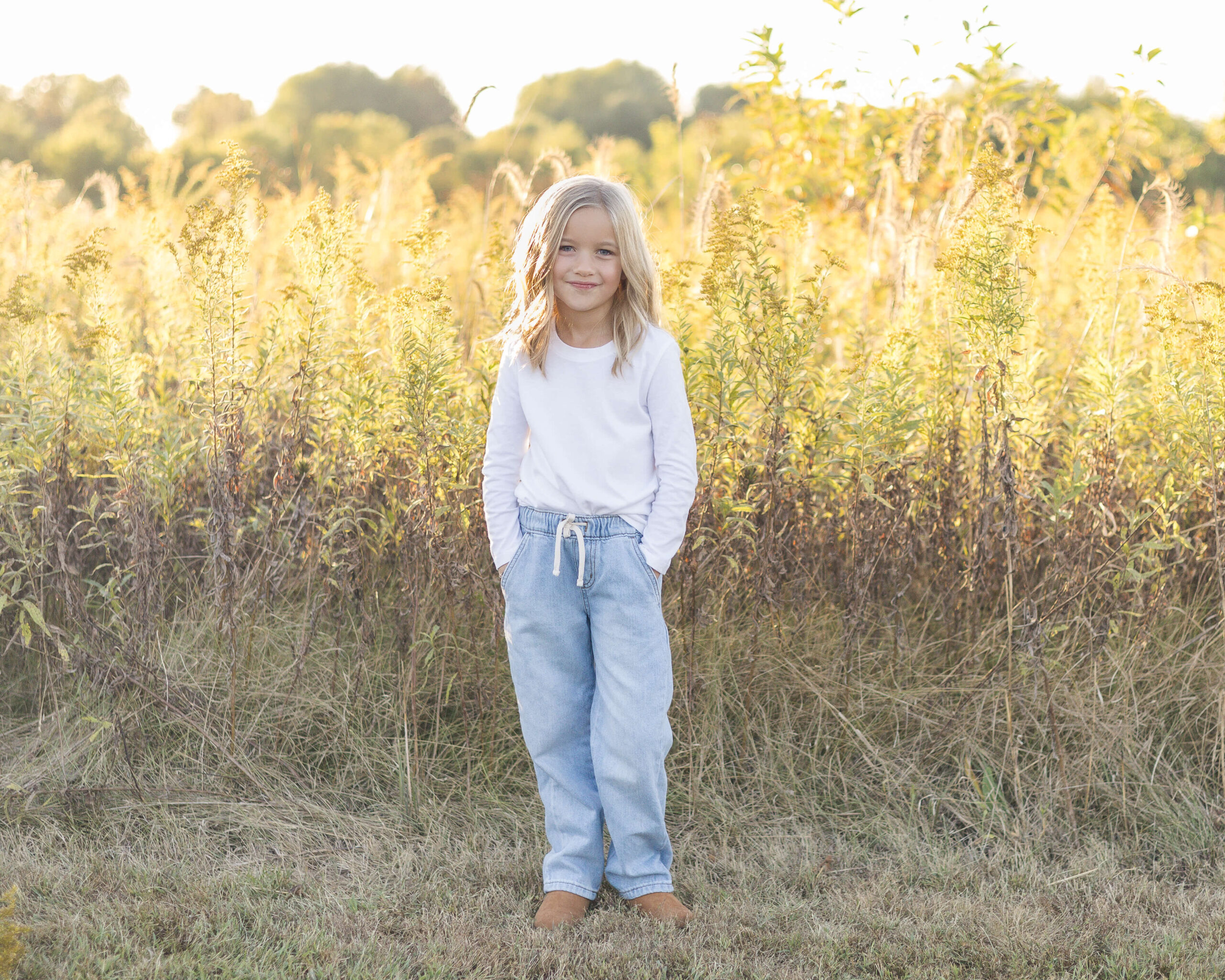 A toddler girl in white and jeans with hands in her pockets in front of tall golden grass at sunset