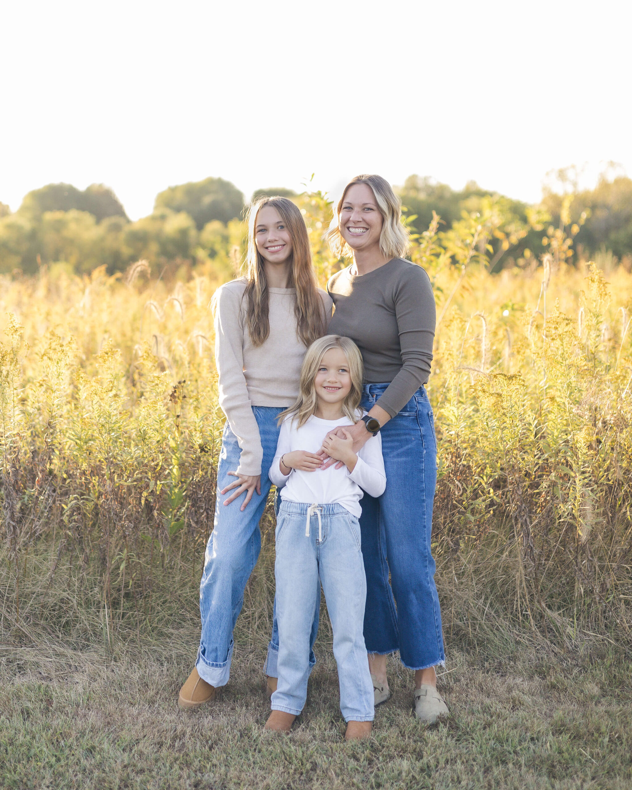 A happy mom stands on the edge of a field at sunset with her two young daughters in jeans and sweaters while enjoying kid-friendly trails in Springfield, IL