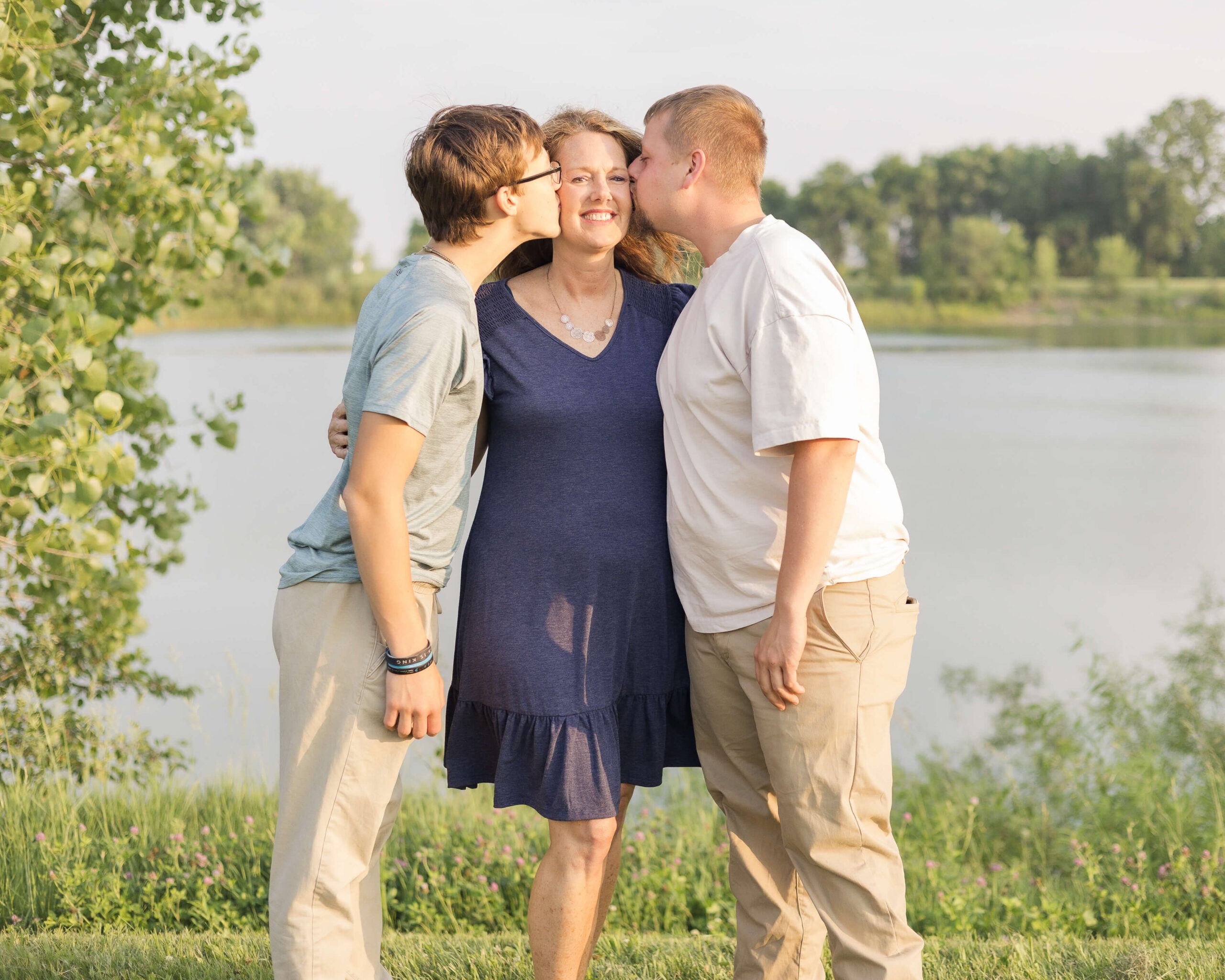 A mom is kissed by her two adult sons on the cheeks by a pond at sunset