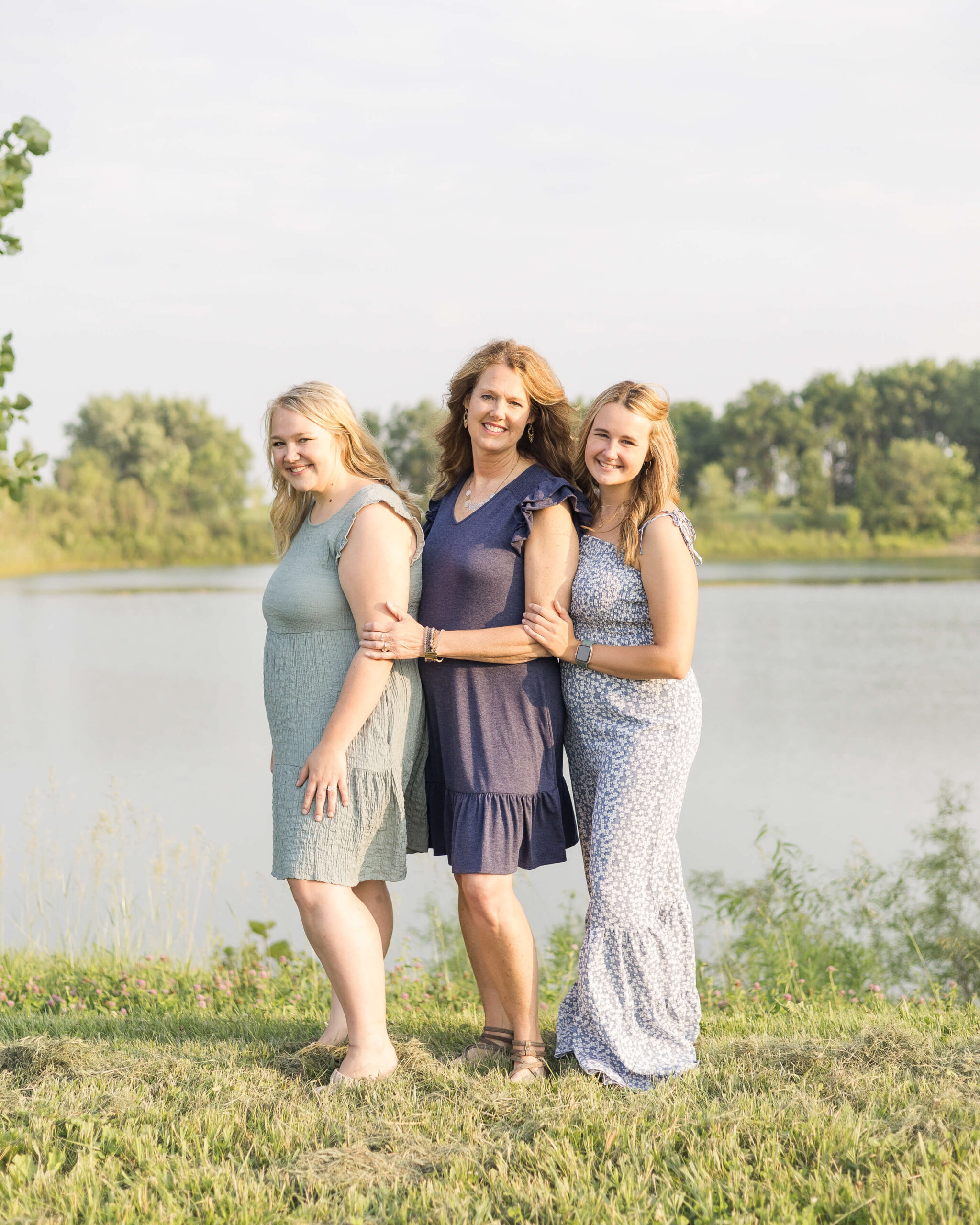 A happy mom in blue dress stands with her two adult daughters by a large pond at sunset after mother's day brunch in springfield il