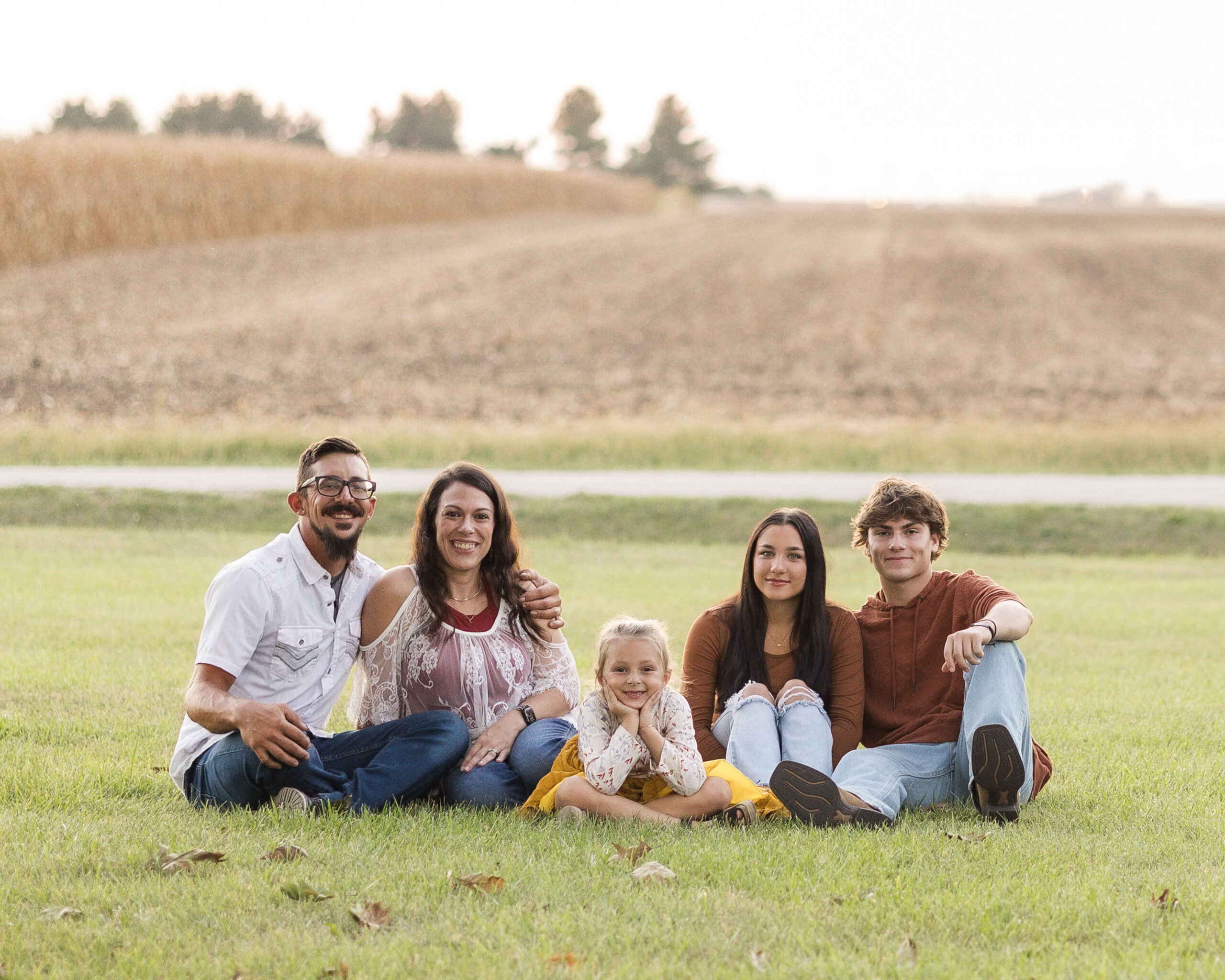 A happy family sits in a grassy lawn by a corn field at sunset smiling