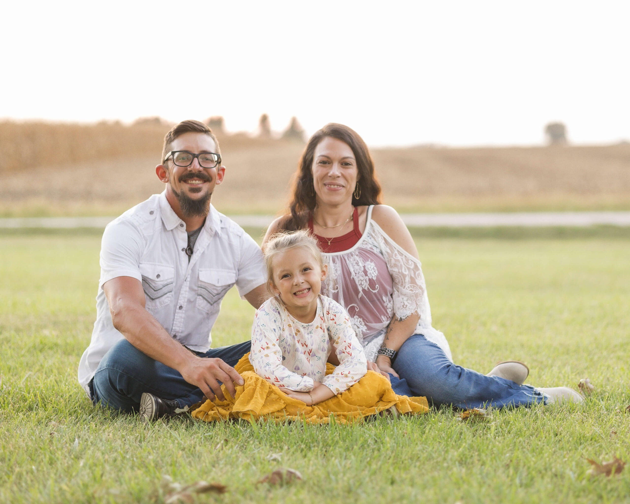 A mom and dad in white sit in a lawn smiling with their toddler daughter in white and yellow at sunset after speech therapy in springfield il