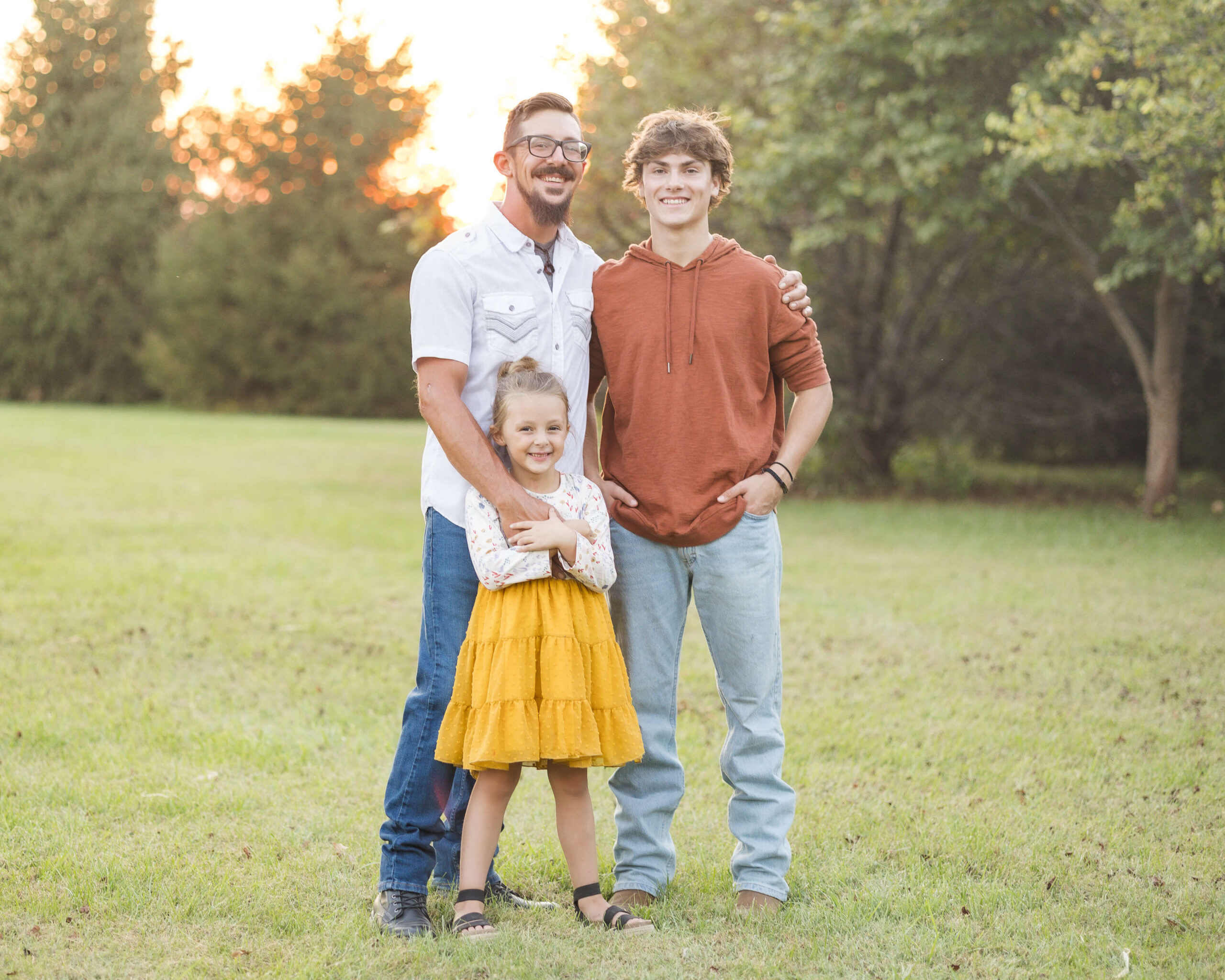 A dad in a whtie shirt stands smiling with his toddler daughter and teen son in a park lawn at sunset after some speech therapy in springfield il