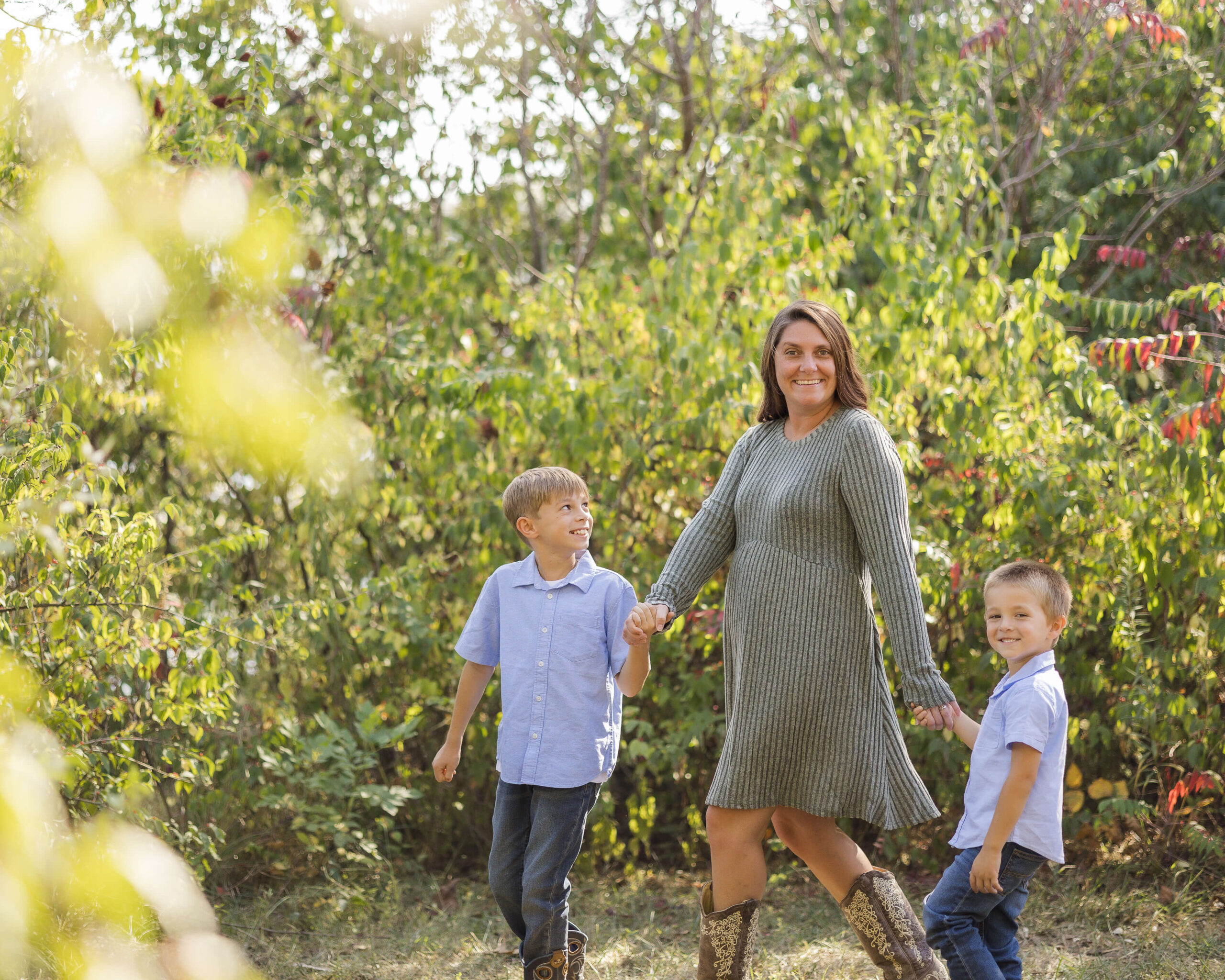 A happy mom in a green dress walks holding hands with her two toddler sons in blue